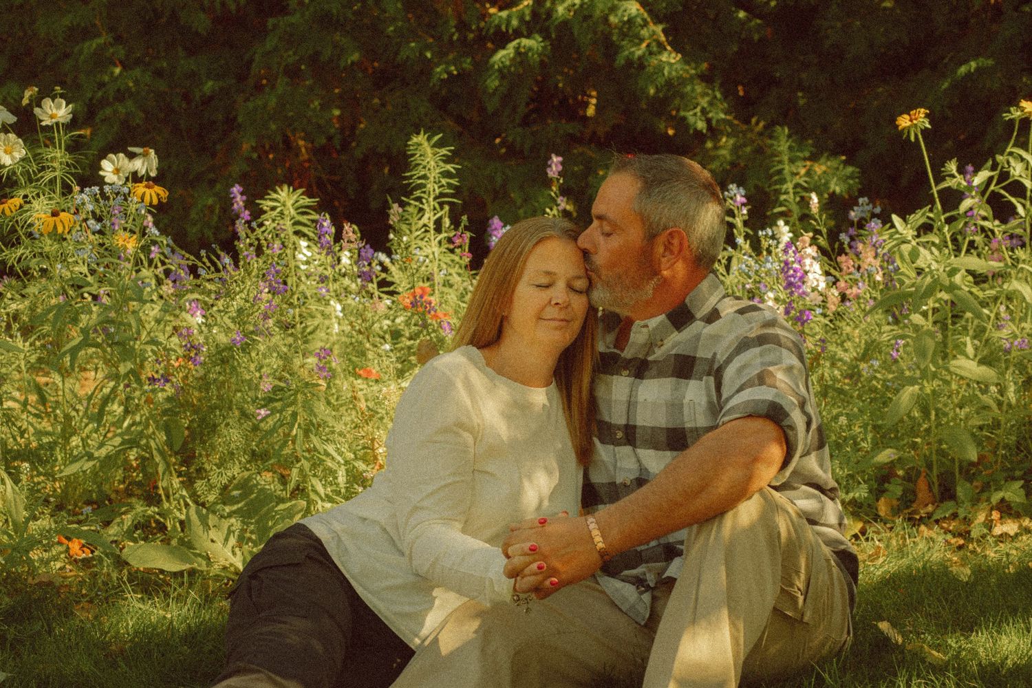 A couple sits in a colorful garden, sharing a tender kiss among blooming wildflowers on a warm sunny day.