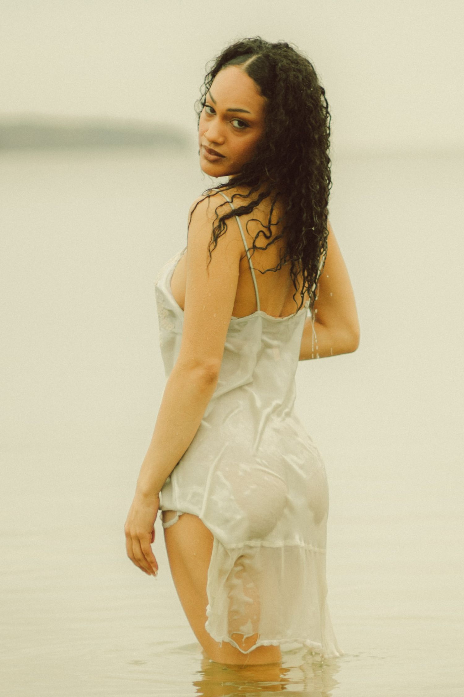 Woman in wet white slip dress standing in shallow water, looking back over shoulder with curly dark hair.