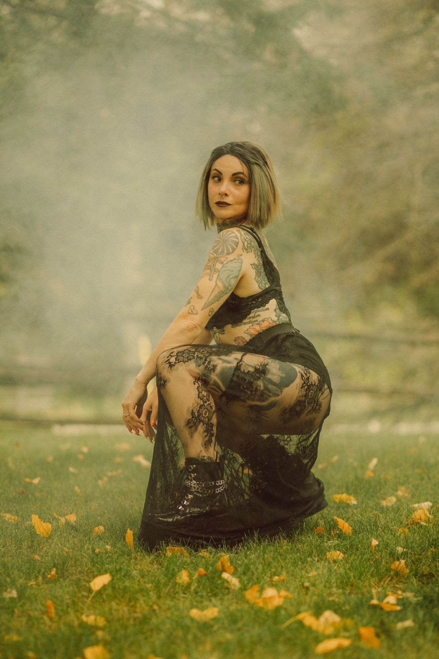 Tattooed woman in black lace dress sitting on grass surrounded by autumn leaves and misty fog in an outdoor setting.