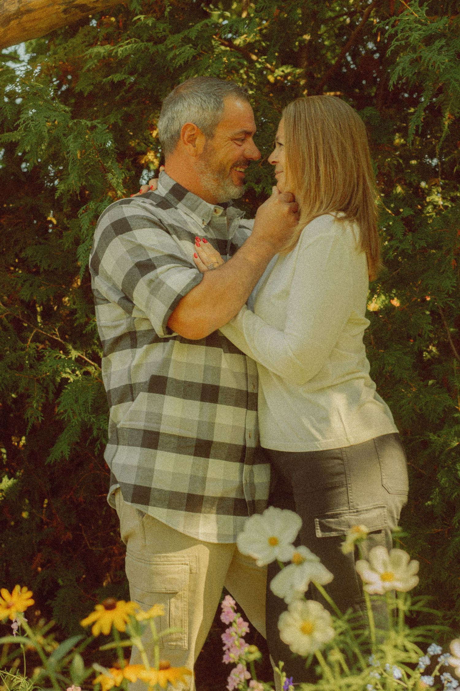 A couple embraces romantically among colorful wildflowers and lush green trees in an outdoor garden setting.