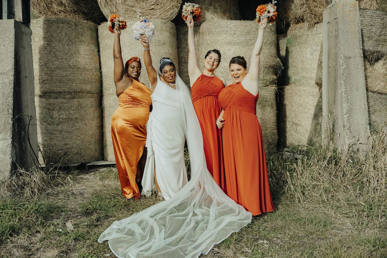 A bridal party poses in orange and white dresses against a rustic stone wall backdrop.