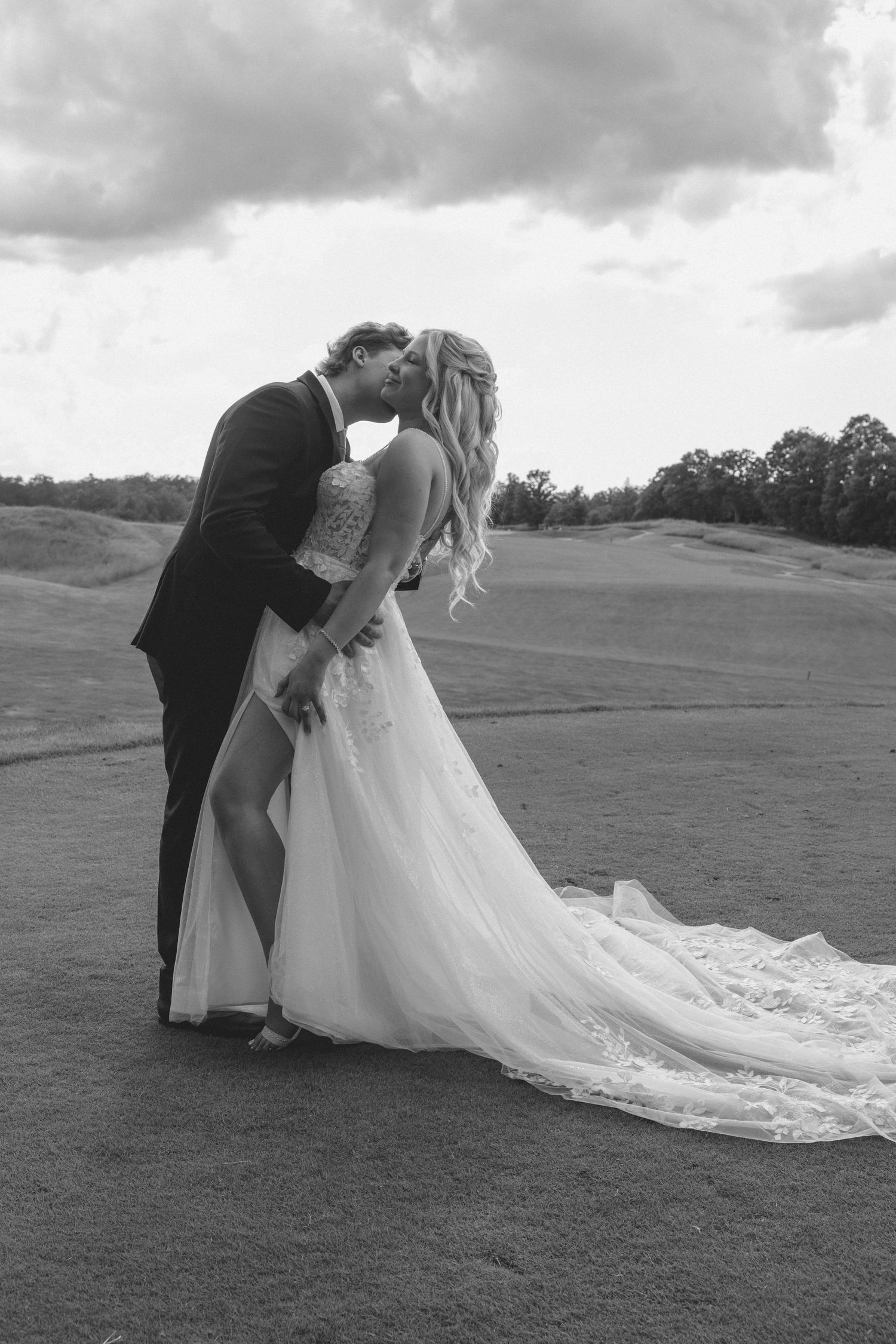 Black and white photo of a bride and groom sharing a romantic kiss outdoors on a golf course on their wedding day.