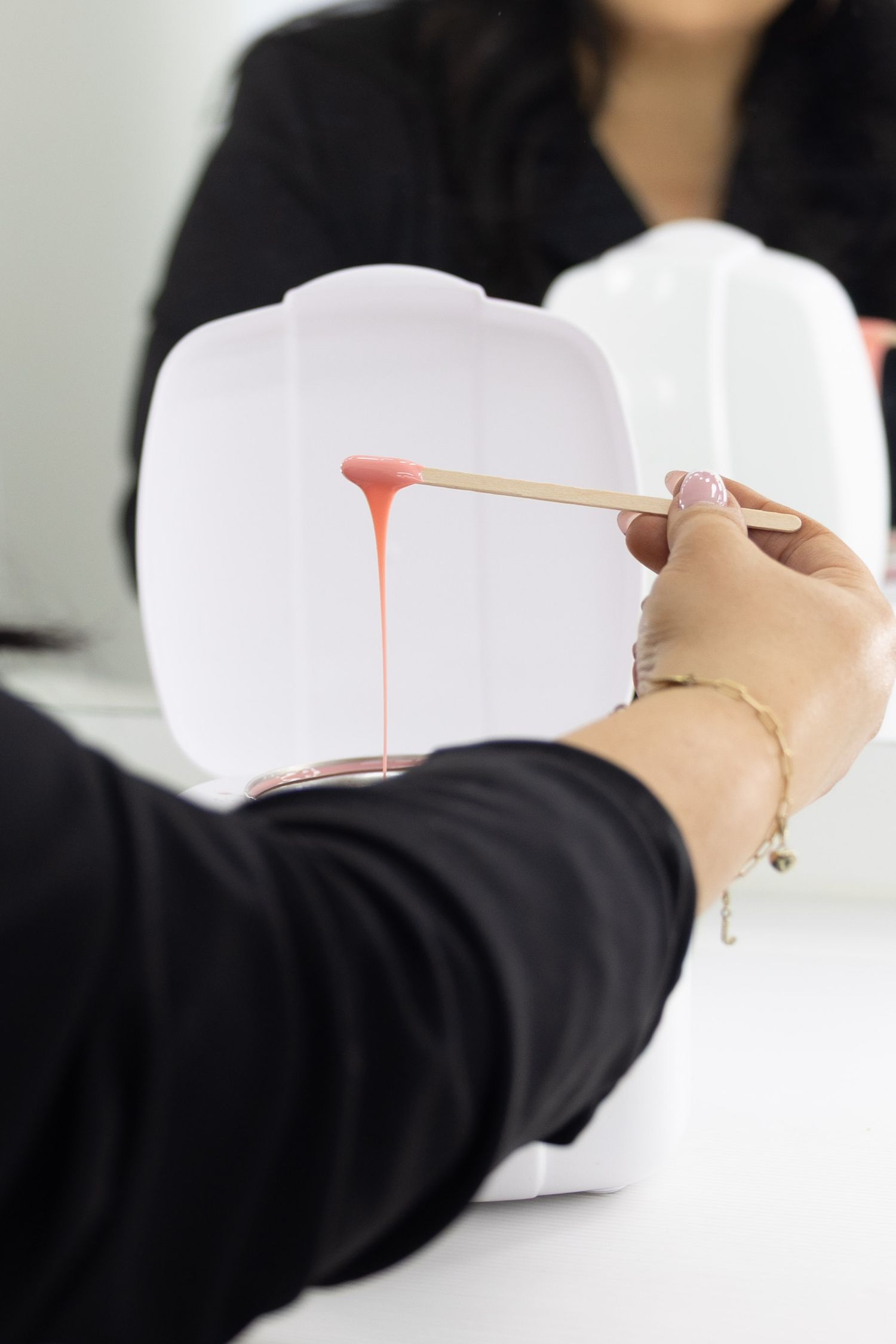 Esthetician holding wooden spatula with pink wax dripping over white wax warmer during hair removal treatment.