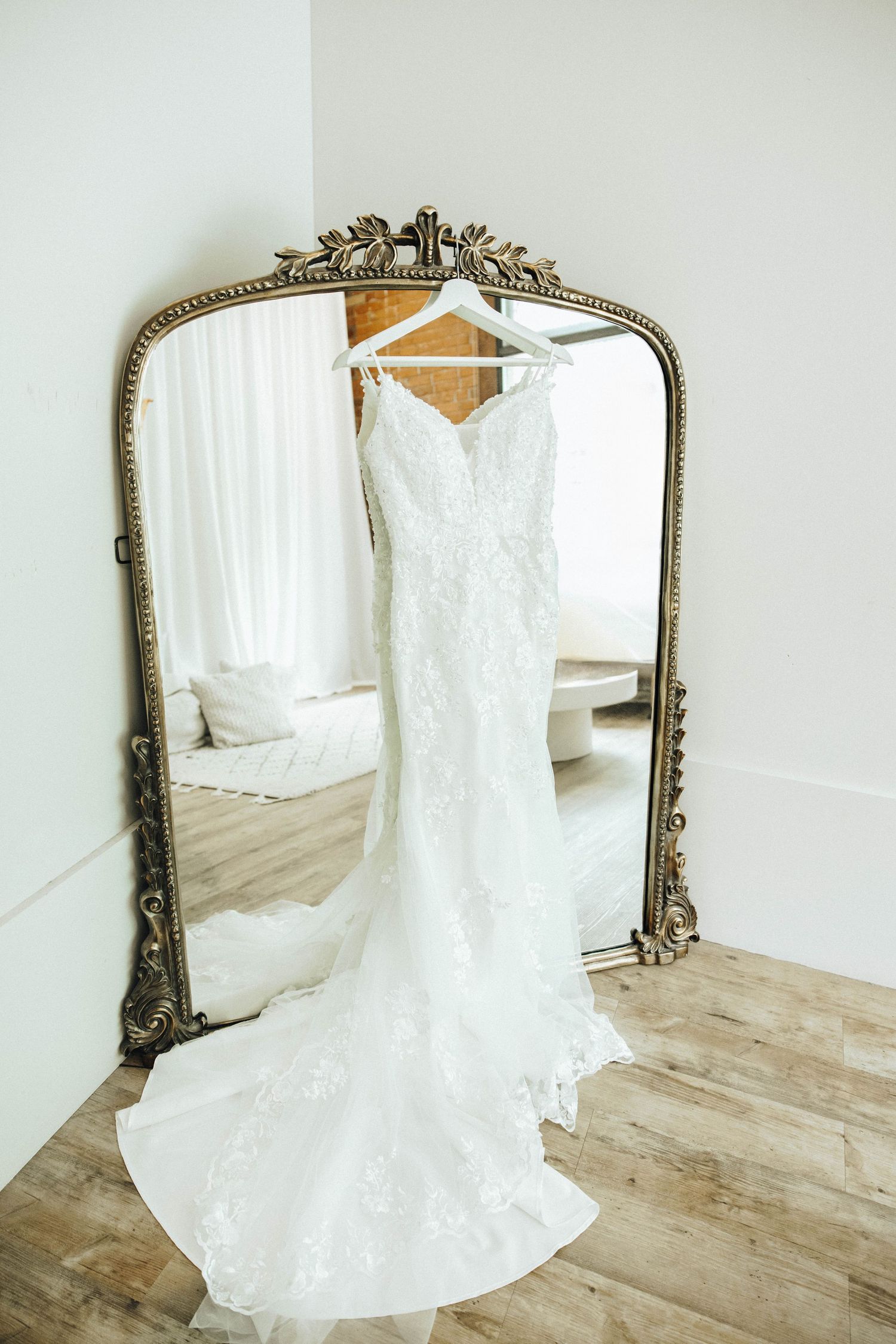 Elegant white lace wedding dress hanging on ornate vintage gold mirror with wooden floor reflection.