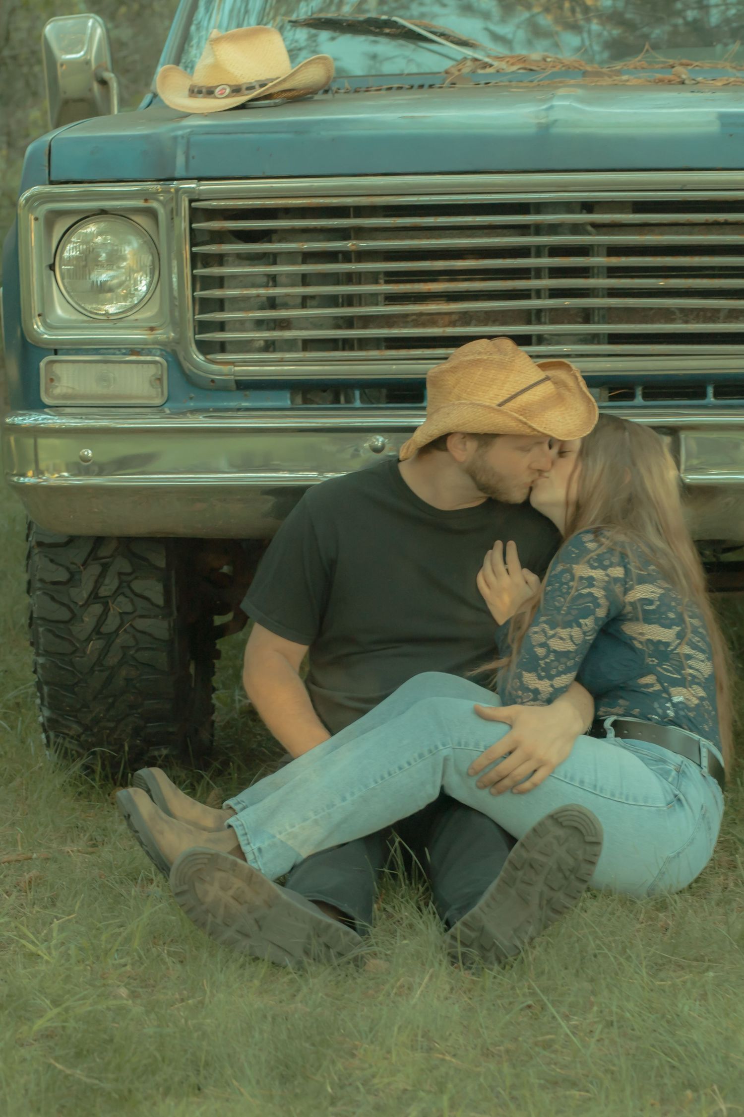 Couple in cowboy hats sharing a kiss while sitting against a vintage blue truck in a rustic outdoor setting.