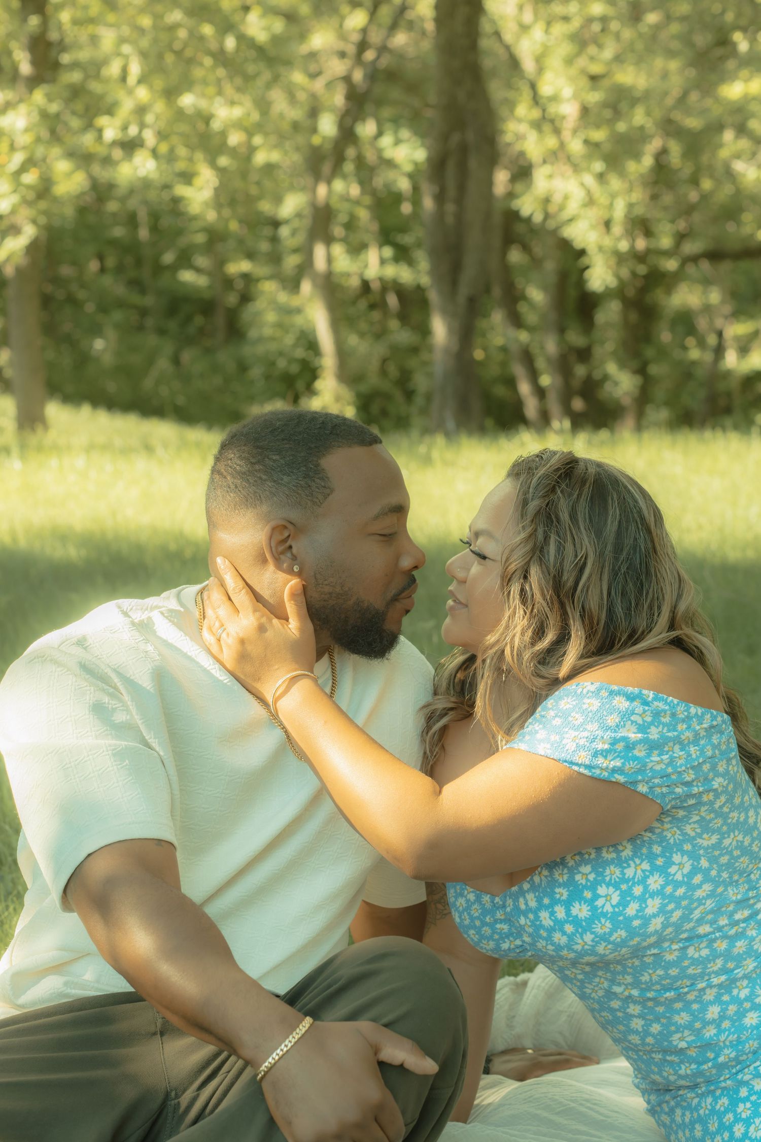 A couple shares a romantic almost-kiss while sitting together on grass in a sunny green park.