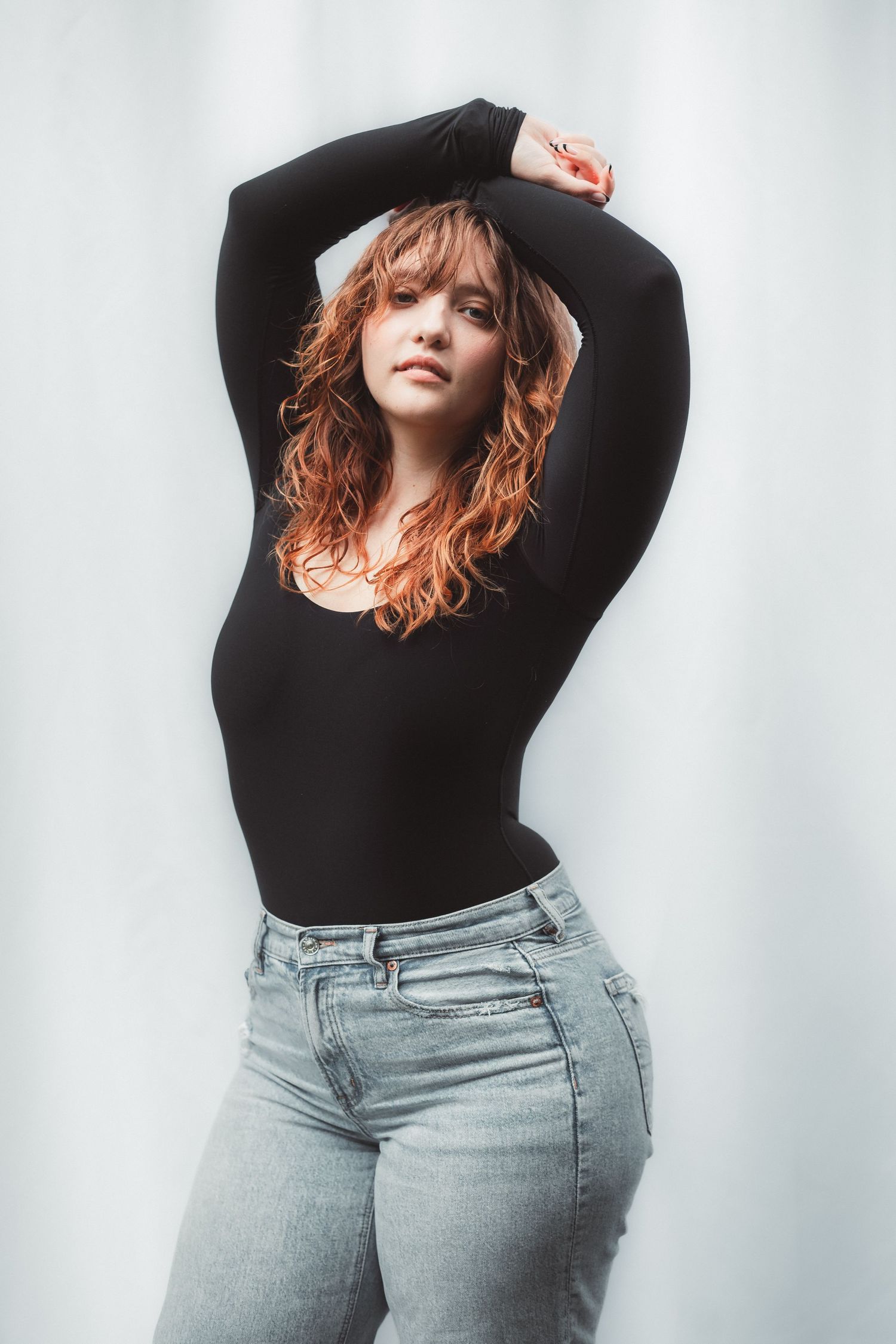 Woman with curly red hair posing in black long-sleeve bodysuit and light wash jeans against white background.