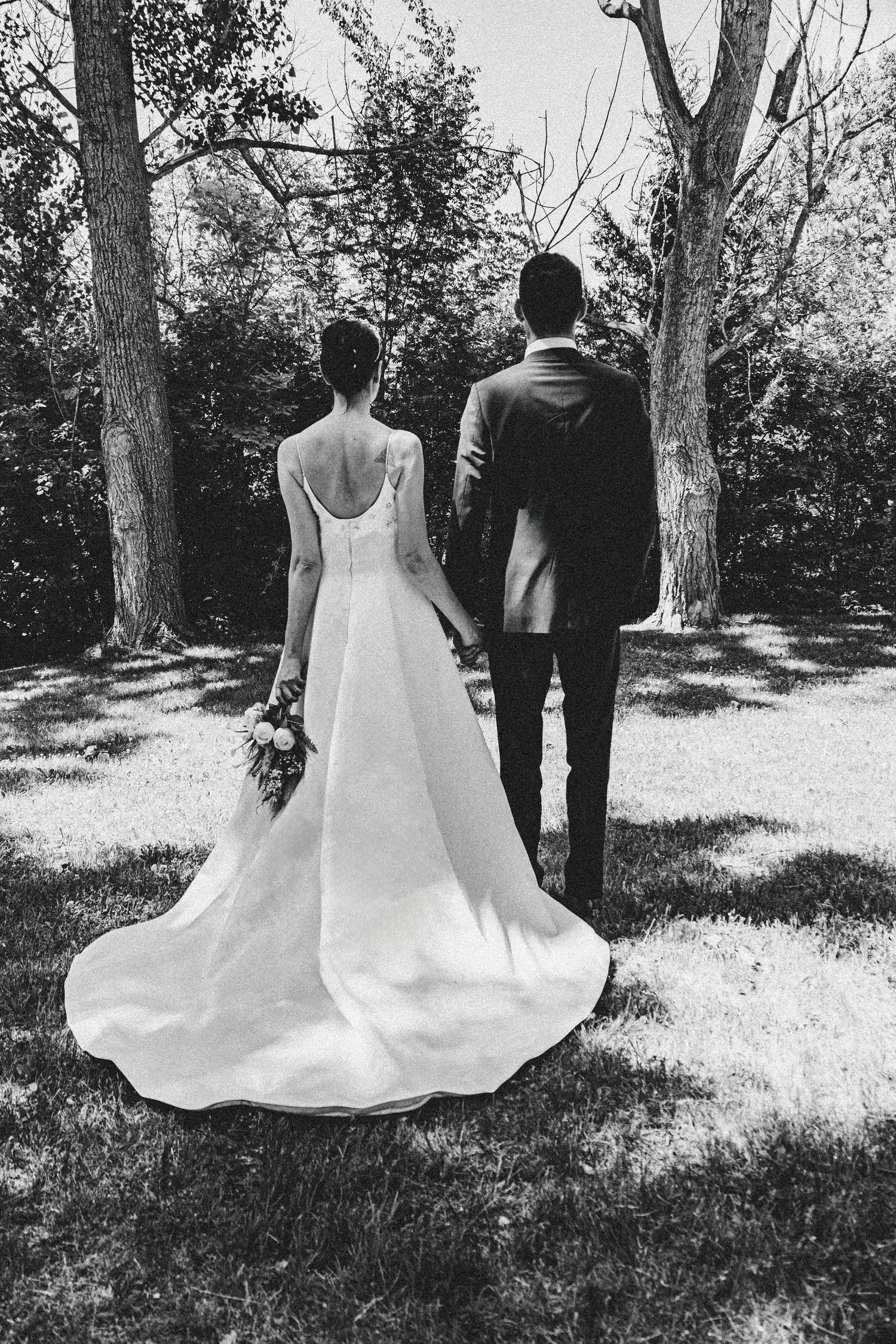 A black and white portrait of newlyweds standing among trees with flowing wedding dress train.