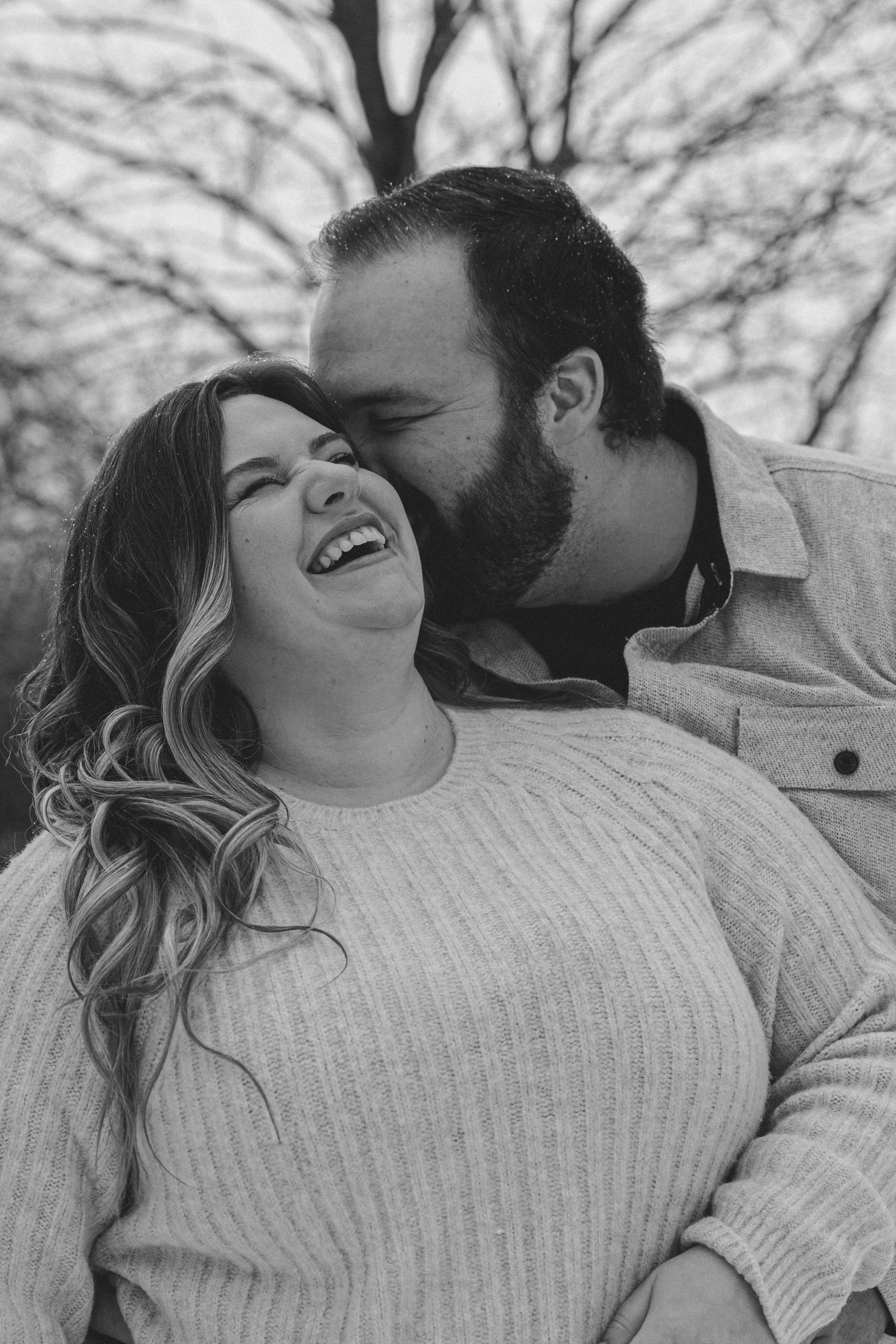 A joyful couple shares an intimate moment outdoors in black and white, with bare tree branches visible in the background.