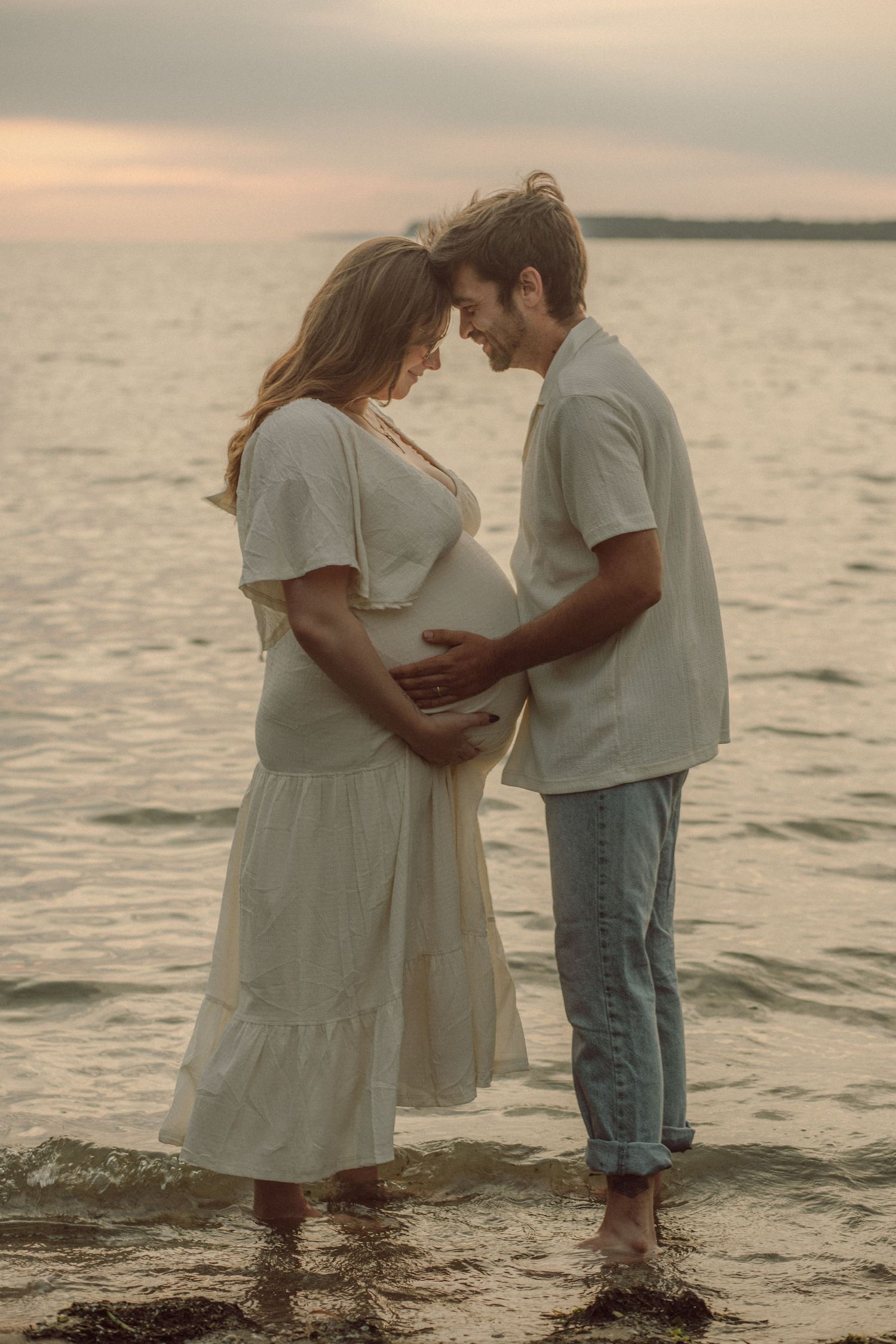 A couple stands barefoot in ocean water at sunset, tenderly touching a pregnant belly in white dress.