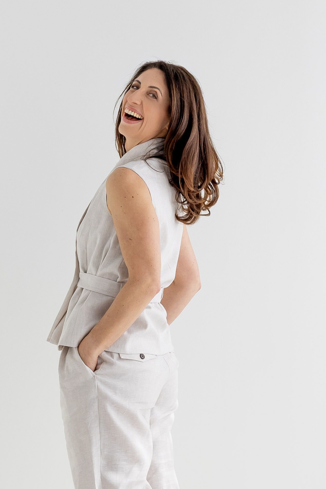 Smiling brunette woman in beige sleeveless top and white pants posing sideways against light background.
