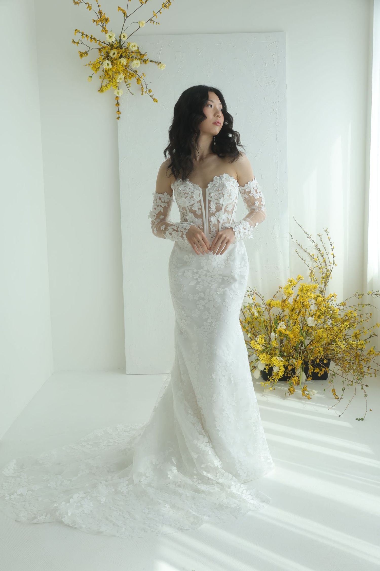 Elegant lace wedding dress with sheer long sleeves and floral details, styled with yellow florals in a bright white setting.