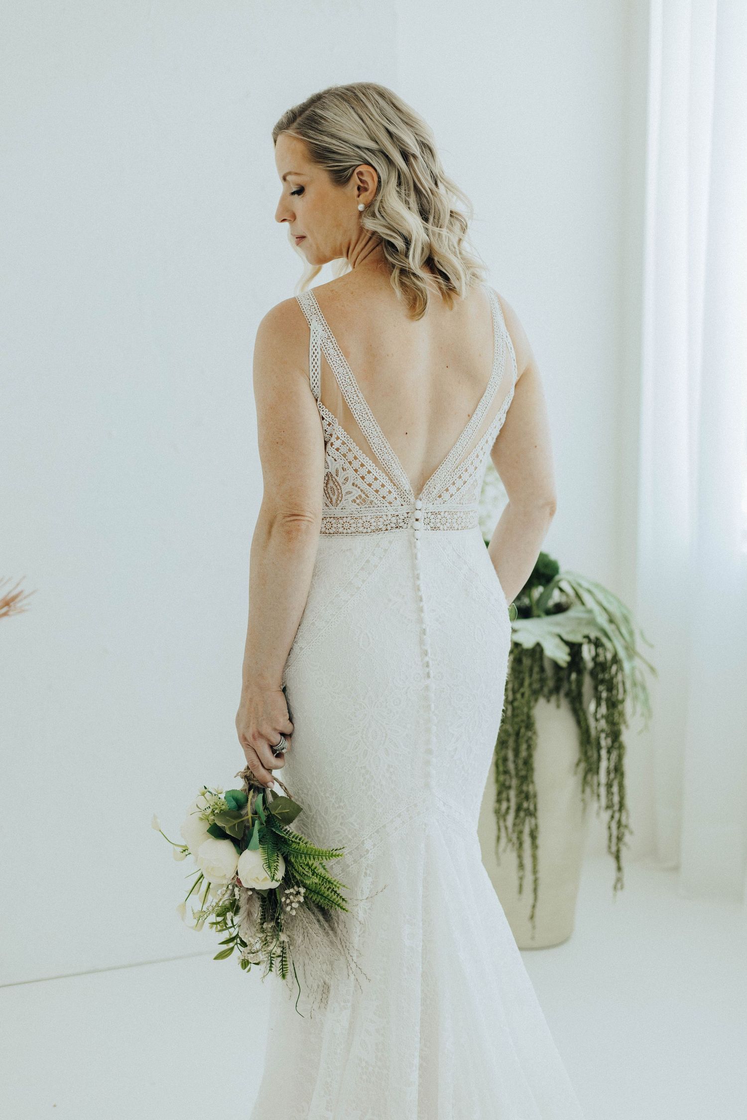 Elegant mermaid wedding dress with lace back detail and trailing greenery bouquet in minimalist white room setting.