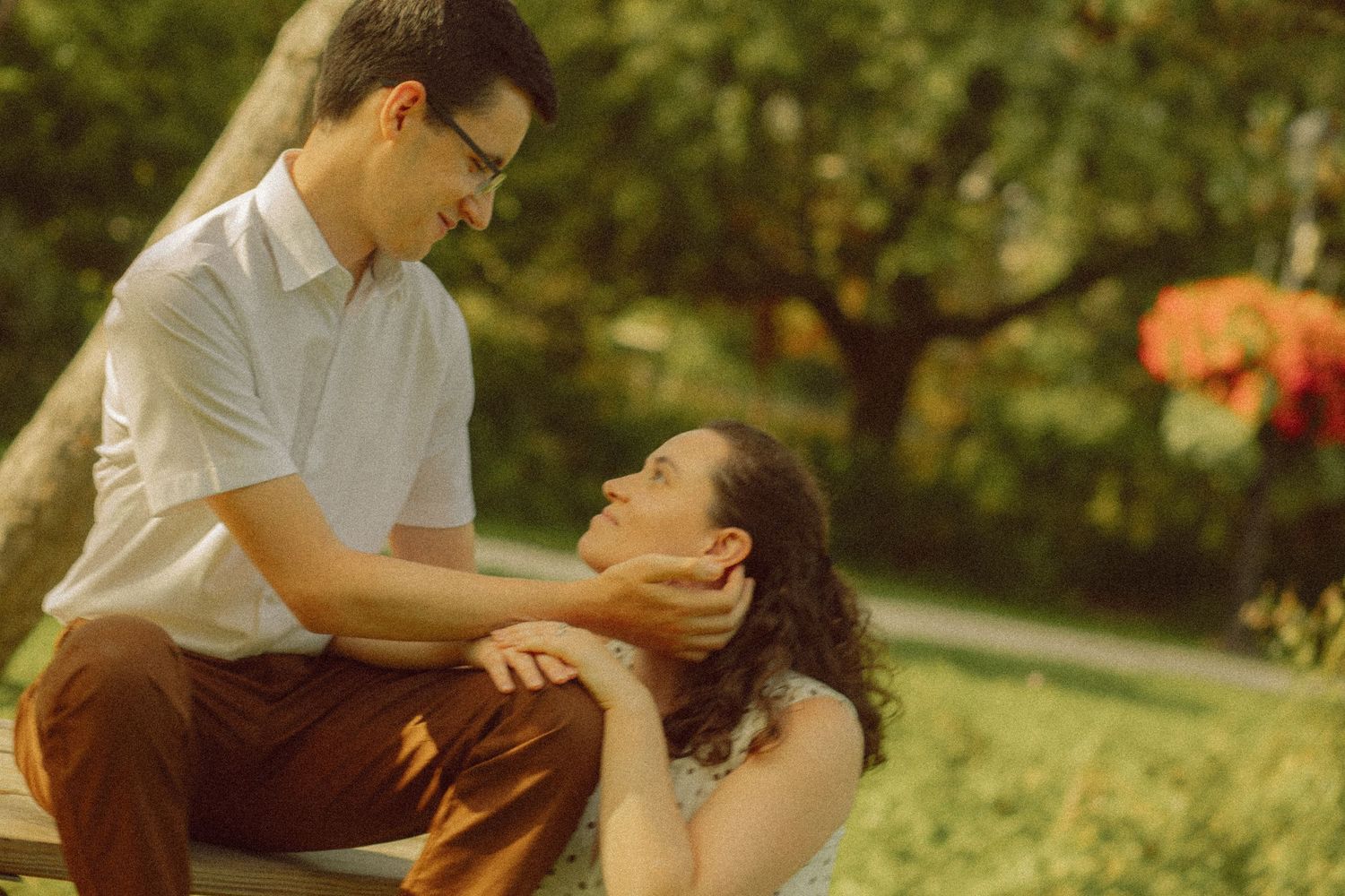 A couple shares a tender moment outdoors, man gently holding womans face while seated on a bench in a lush garden.