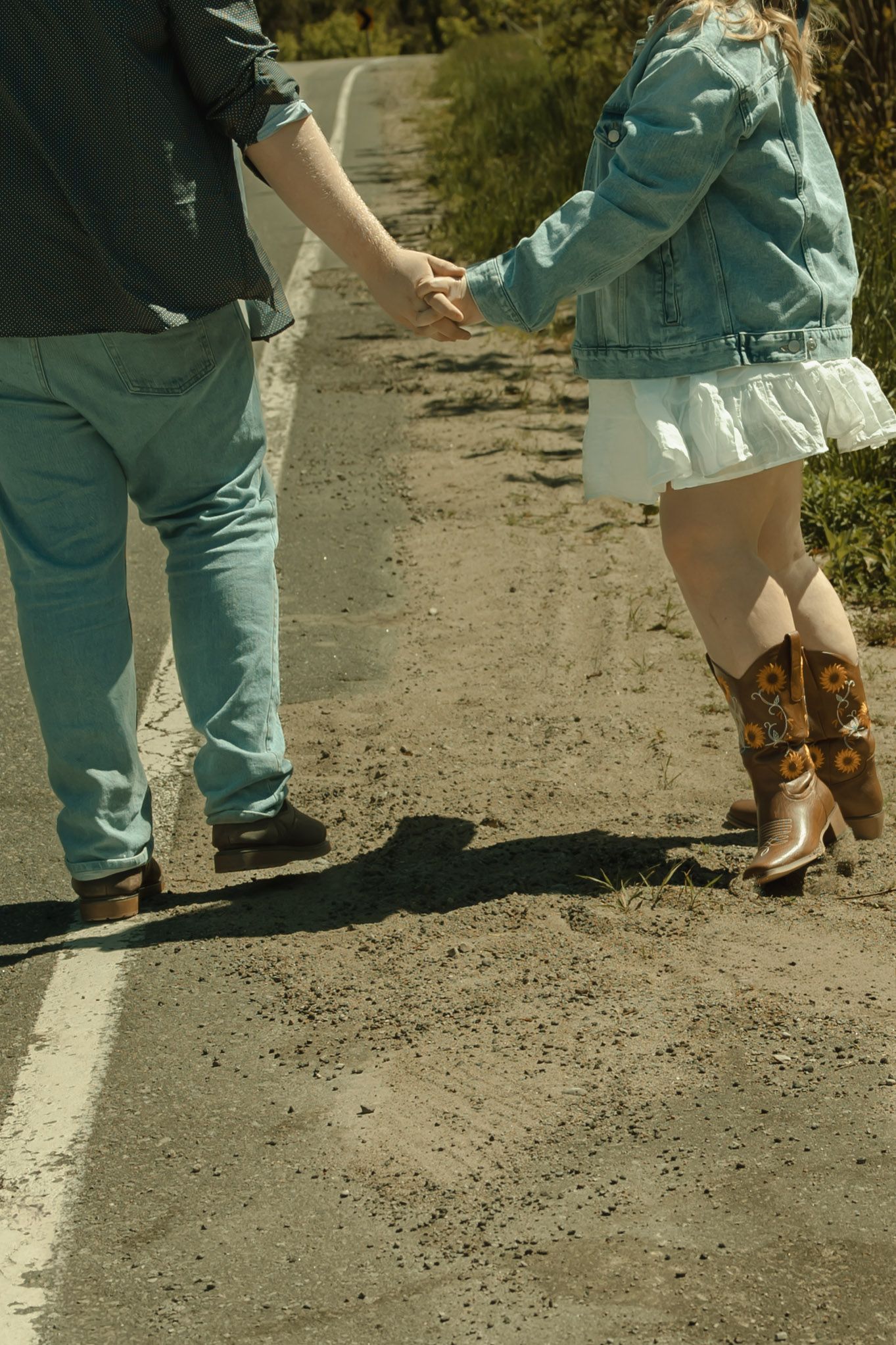 A couple holds hands while walking on a dirt road, wearing denim and cowboy boots in a rural outdoor setting.