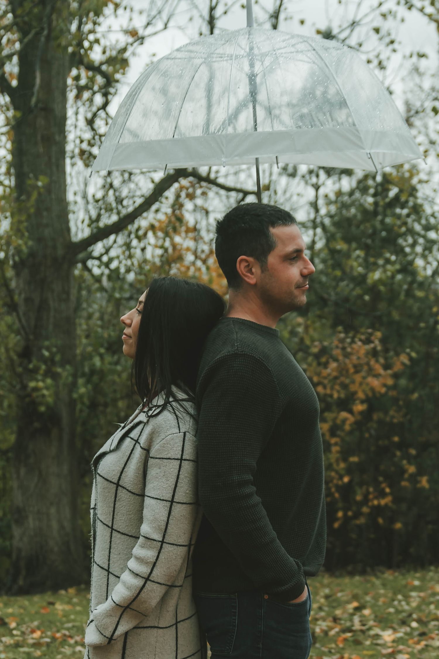 A couple stands back to back outdoors in an autumn setting with fallen leaves and trees in the background.