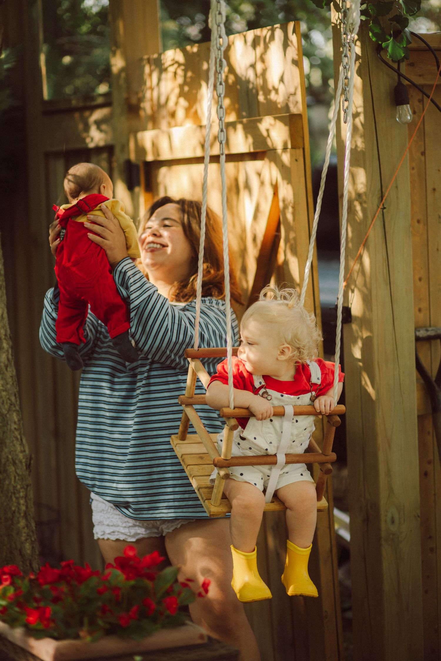 Mother holding baby in red outfit beside toddler on wooden swing in sunny garden setting.