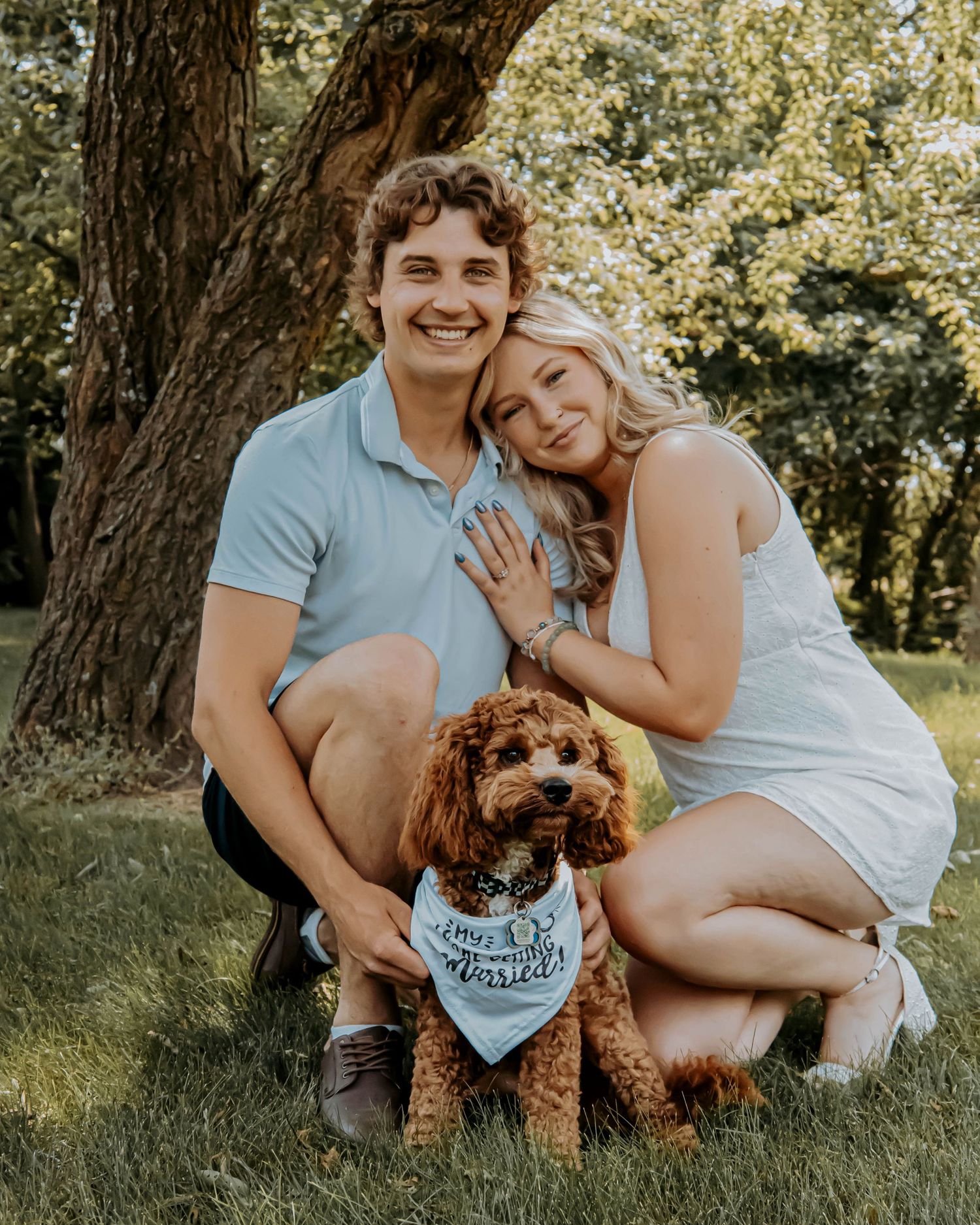 A couple sits on grass with their adorable brown toy poodle during a sunny outdoor photo session.