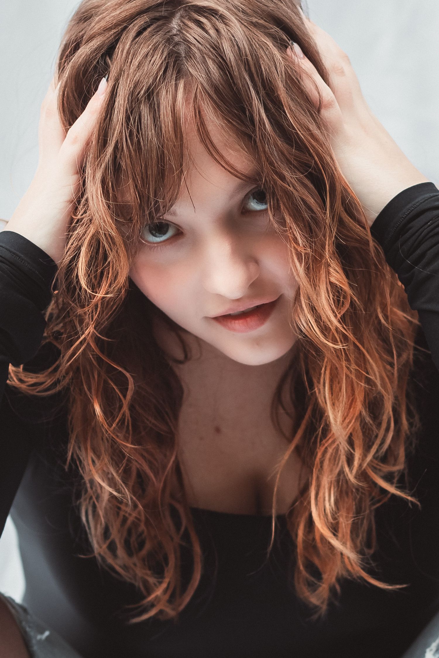 Young woman with long wavy auburn hair and blue eyes posing in a black top, hands in hair.