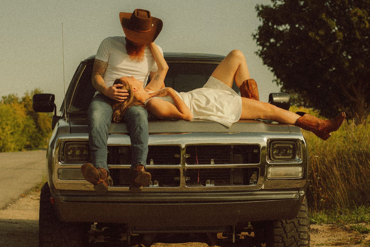 Couple in cowboy hats relaxing on vintage truck hood on rural road, country lifestyle photography.
