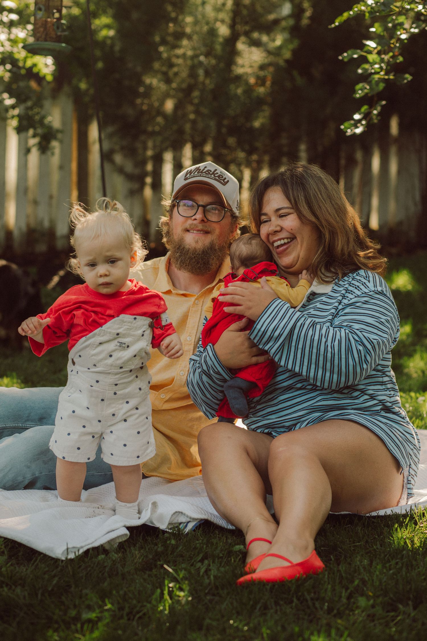 A happy family of four sits on a blanket in a sunny backyard, with two young children in colorful outfits.