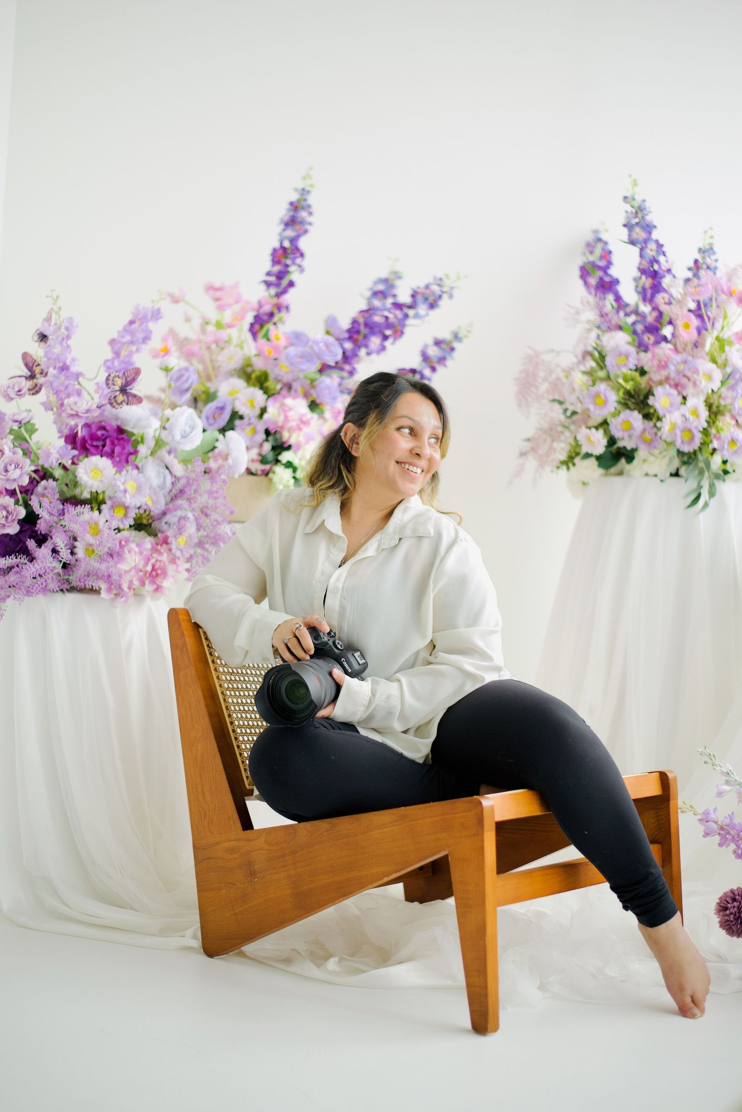 Woman photographer sitting in wooden chair holding camera surrounded by purple and pink floral arrangements in bright studio.