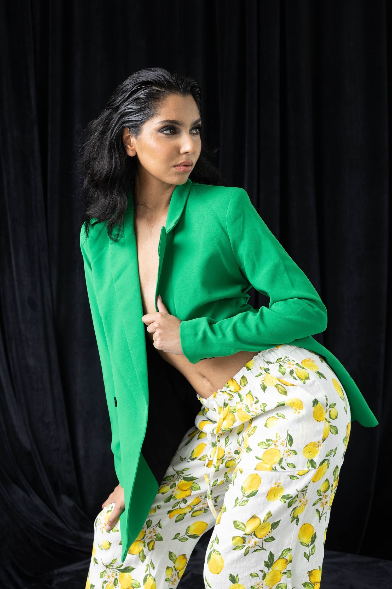 Fashion model poses in bright green blazer and lemon print pants against black backdrop.