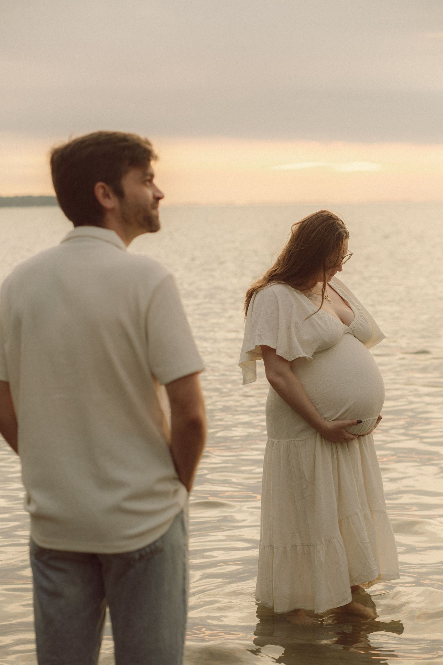 A pregnant woman in a white dress cradles her belly at sunset by the water as a man watches from behind.