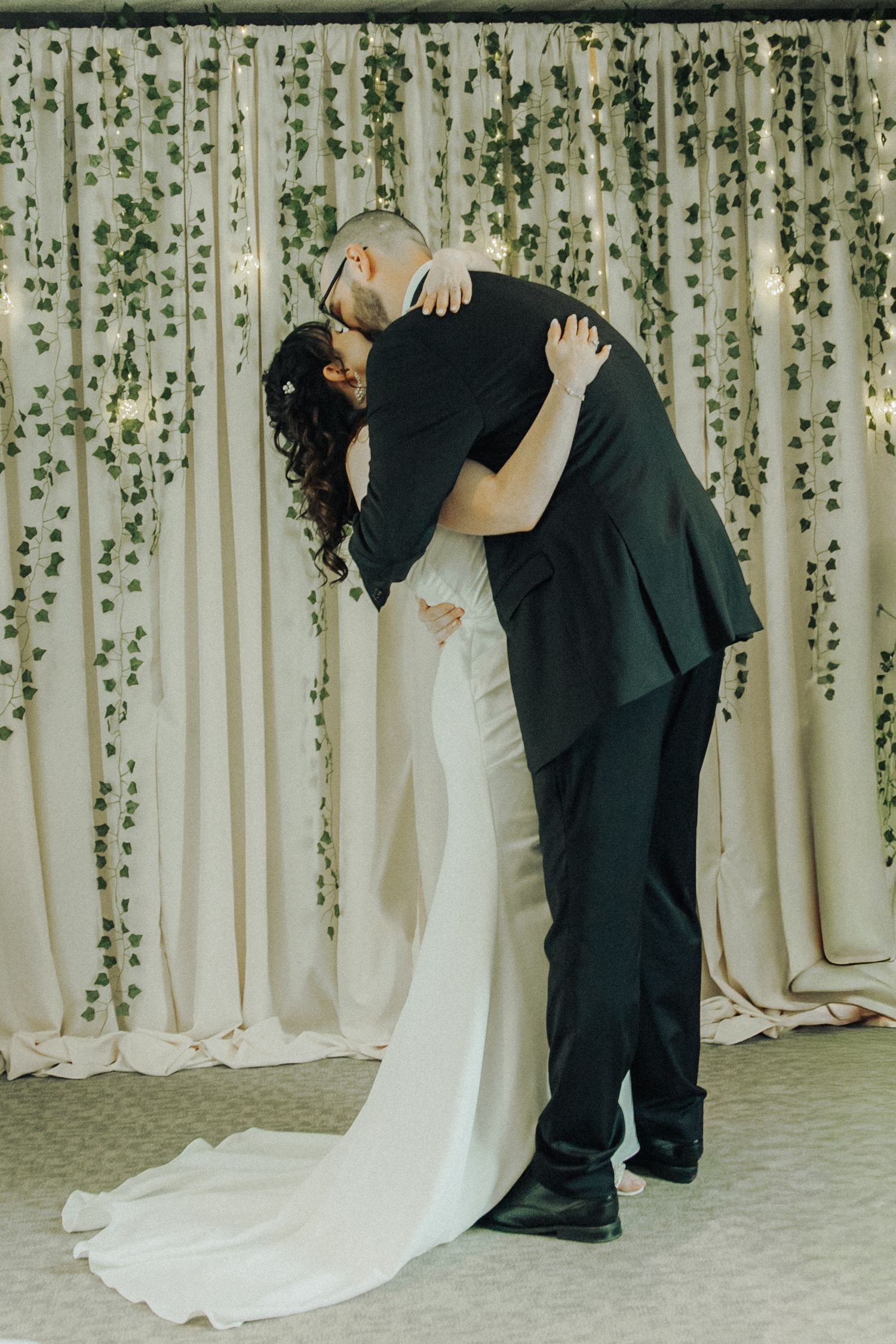Bride and groom sharing first kiss at altar with elegant ivy and fairy light backdrop.