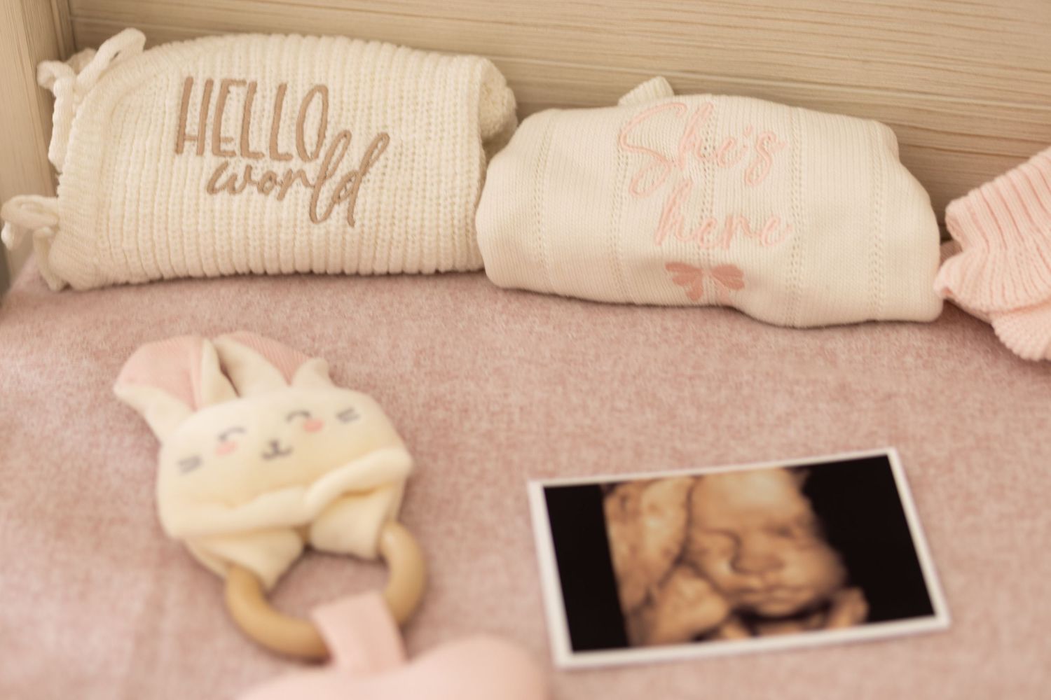 Baby nursery flat lay with ultrasound photo, bunny rattle, and Hello World pillow on pink bedding.