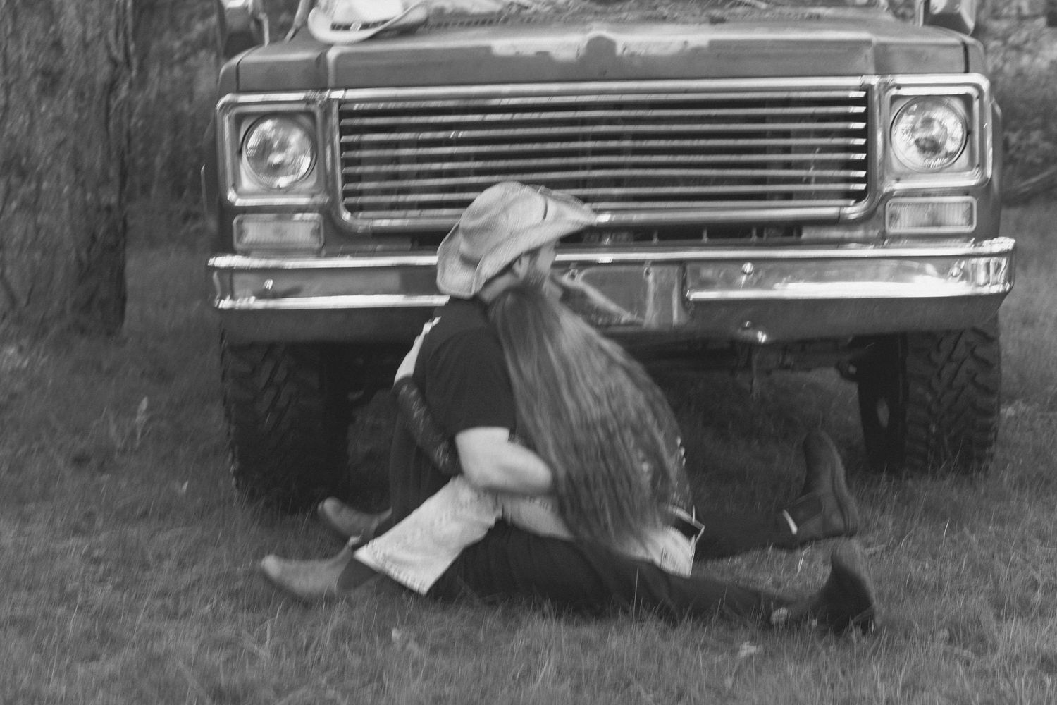 Black and white photo of two people embracing near a vintage truck parked by a tree on grass.