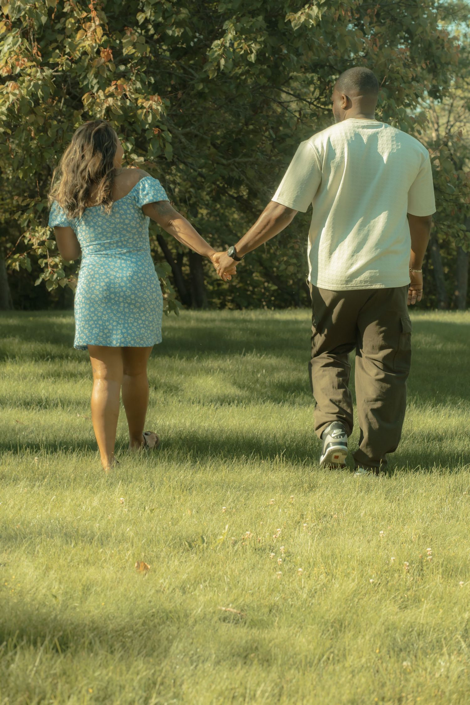 Couple holding hands walking through green park, woman in blue dress and man in white shirt, rear view.