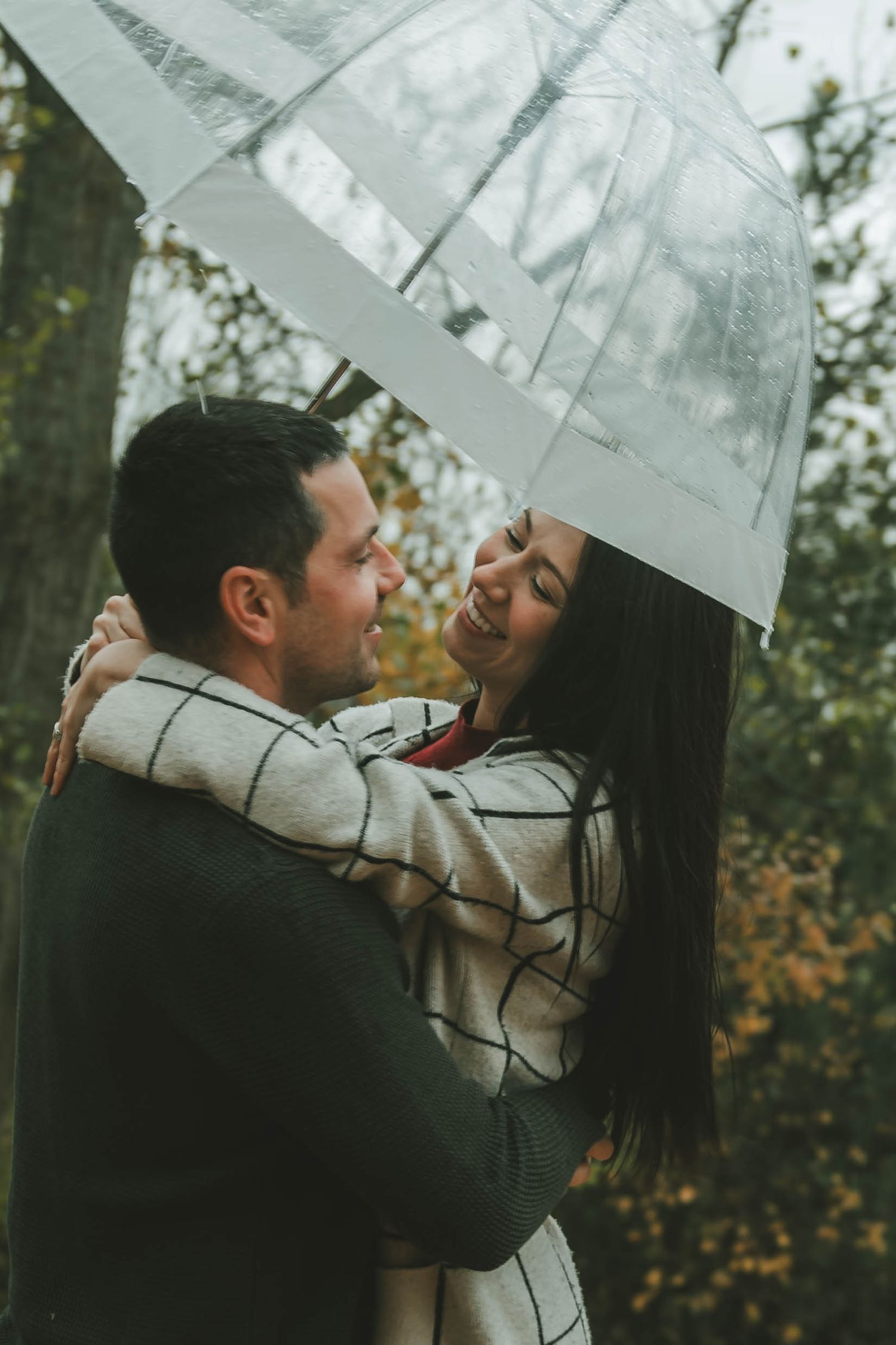 A couple embraces under a clear umbrella surrounded by fall foliage in a park.