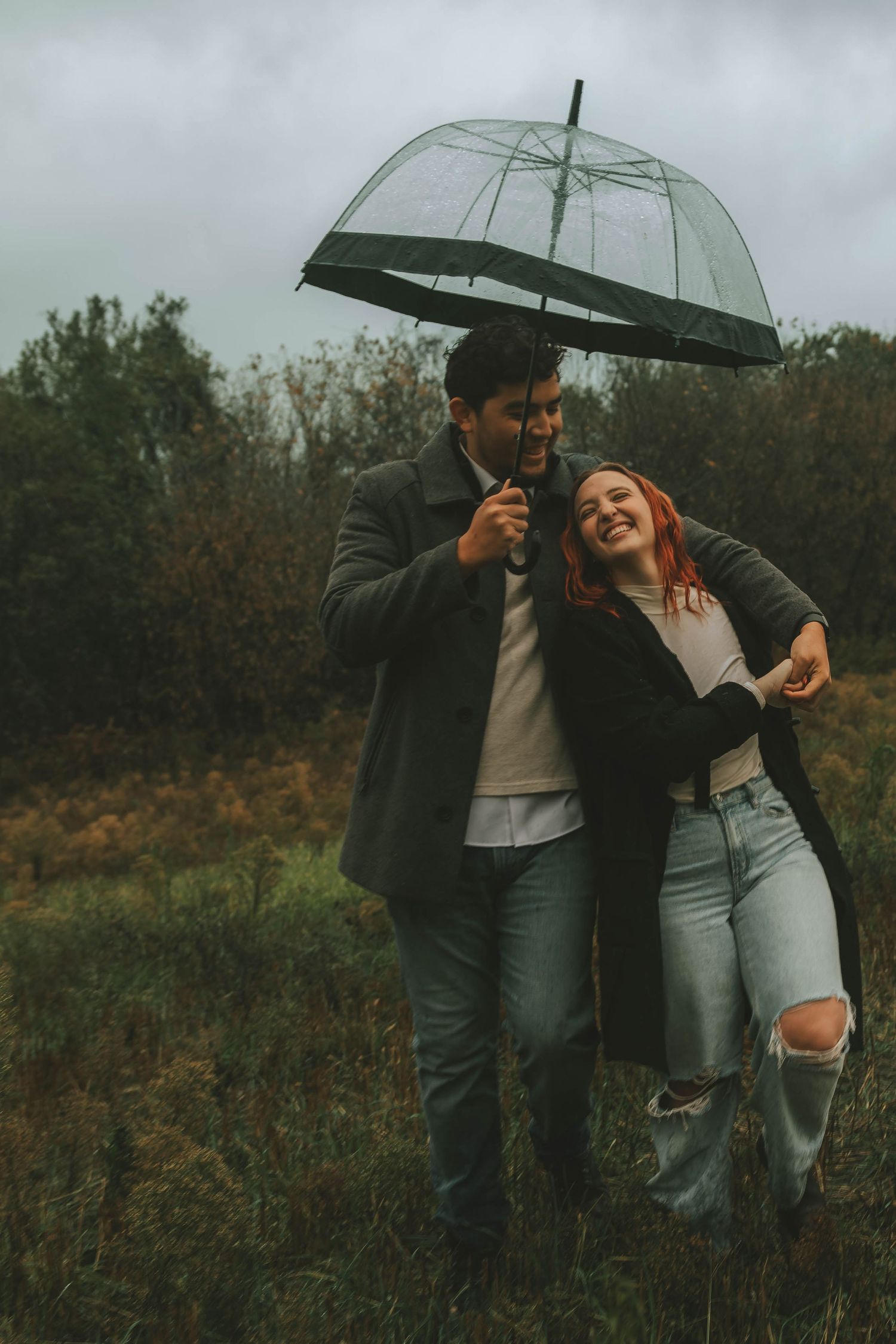 Couple embracing under transparent umbrella on overcast fall day in field.