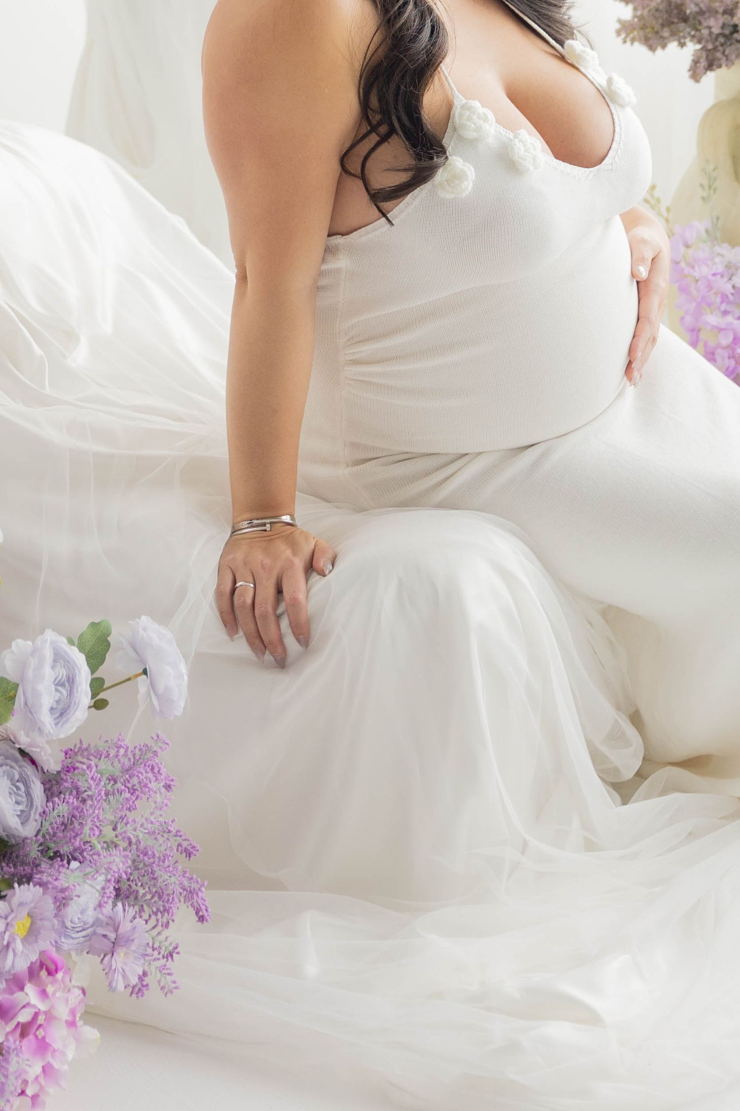 Elegant white maternity gown with flowing tulle fabric and delicate purple flowers in soft natural lighting.
