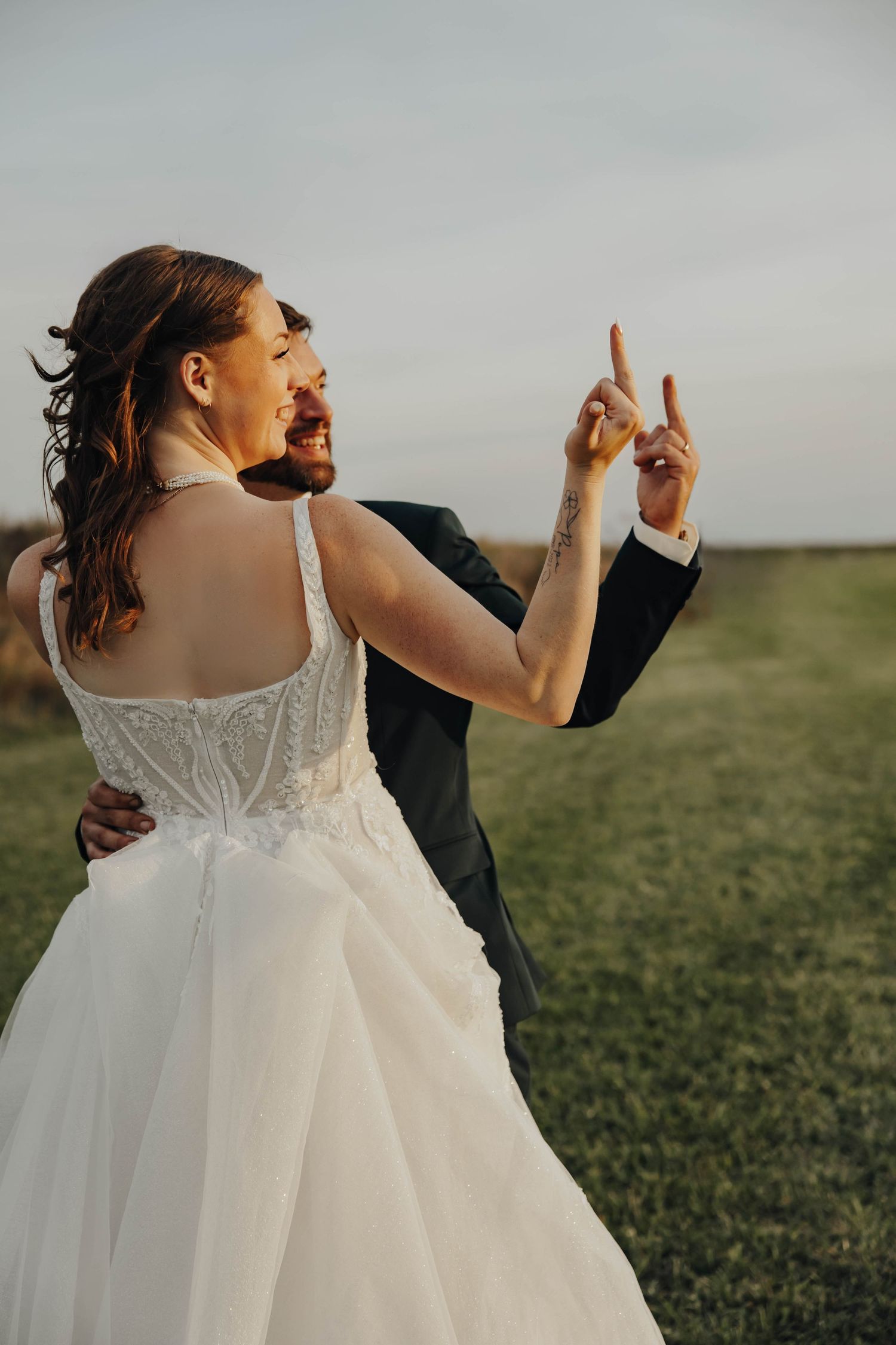 A couple in wedding attire dances playfully in a grassy field during golden hour.
