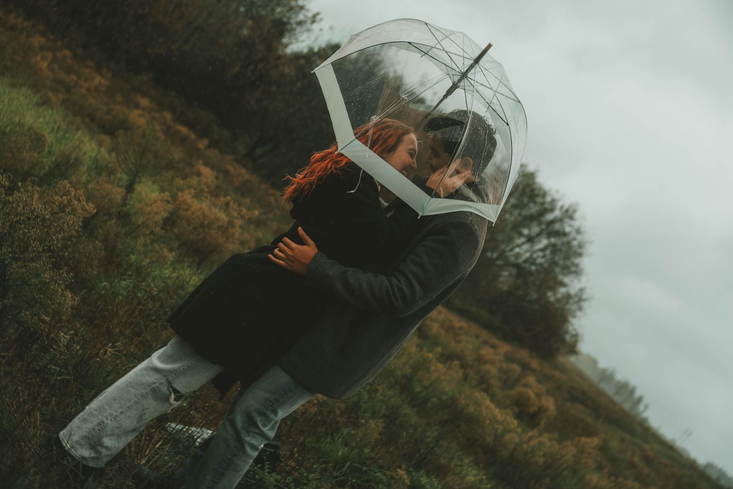 Couple laughing together while holding clear umbrella in moody autumn landscape.