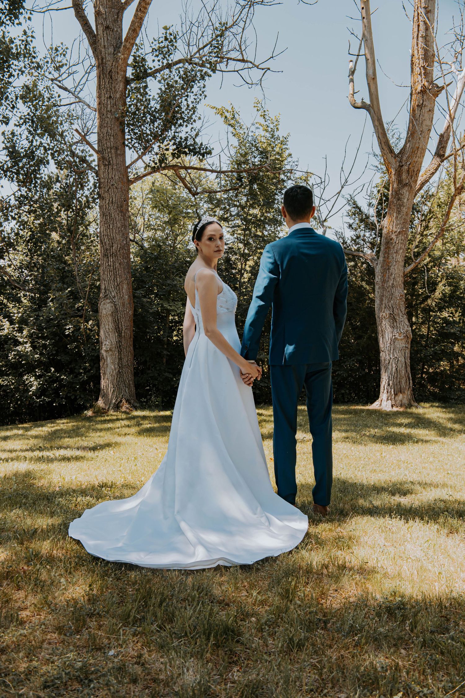 A couple in wedding attire stands hand-in-hand among tall trees in natural sunlight.