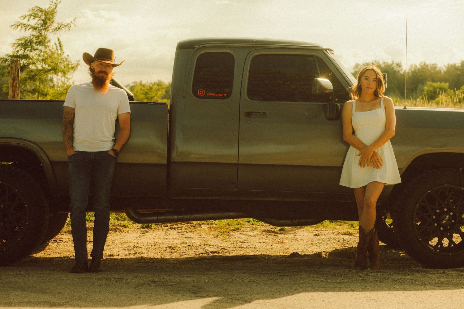 A man in a cowboy hat and a woman in a white dress stand beside a gray pickup truck outdoors.