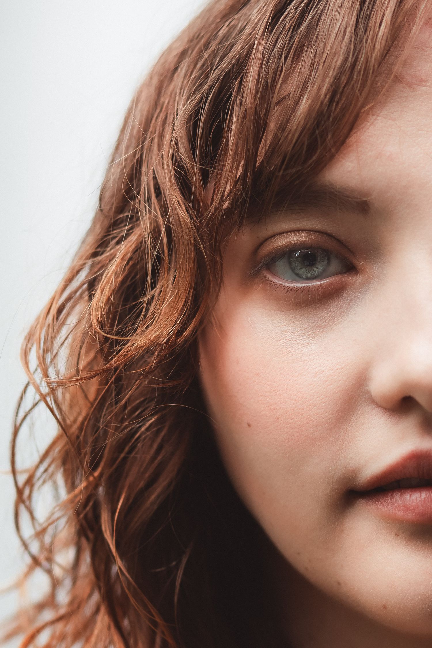 Close-up portrait of a young woman with auburn wavy hair and blue-gray eyes against a light background.