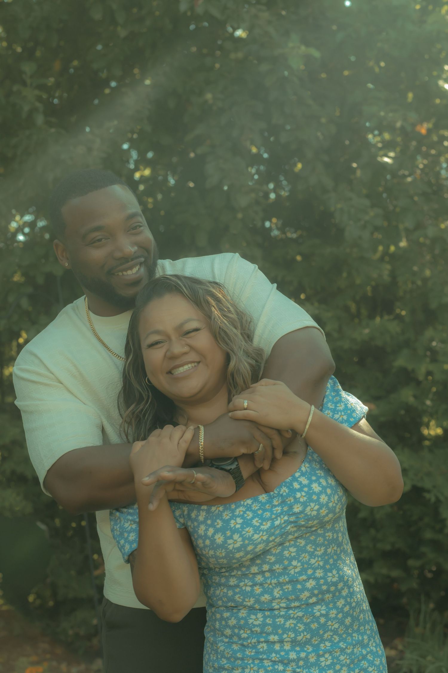 A joyful couple embracing in a sunlit park, the man hugging the woman from behind as they smile.