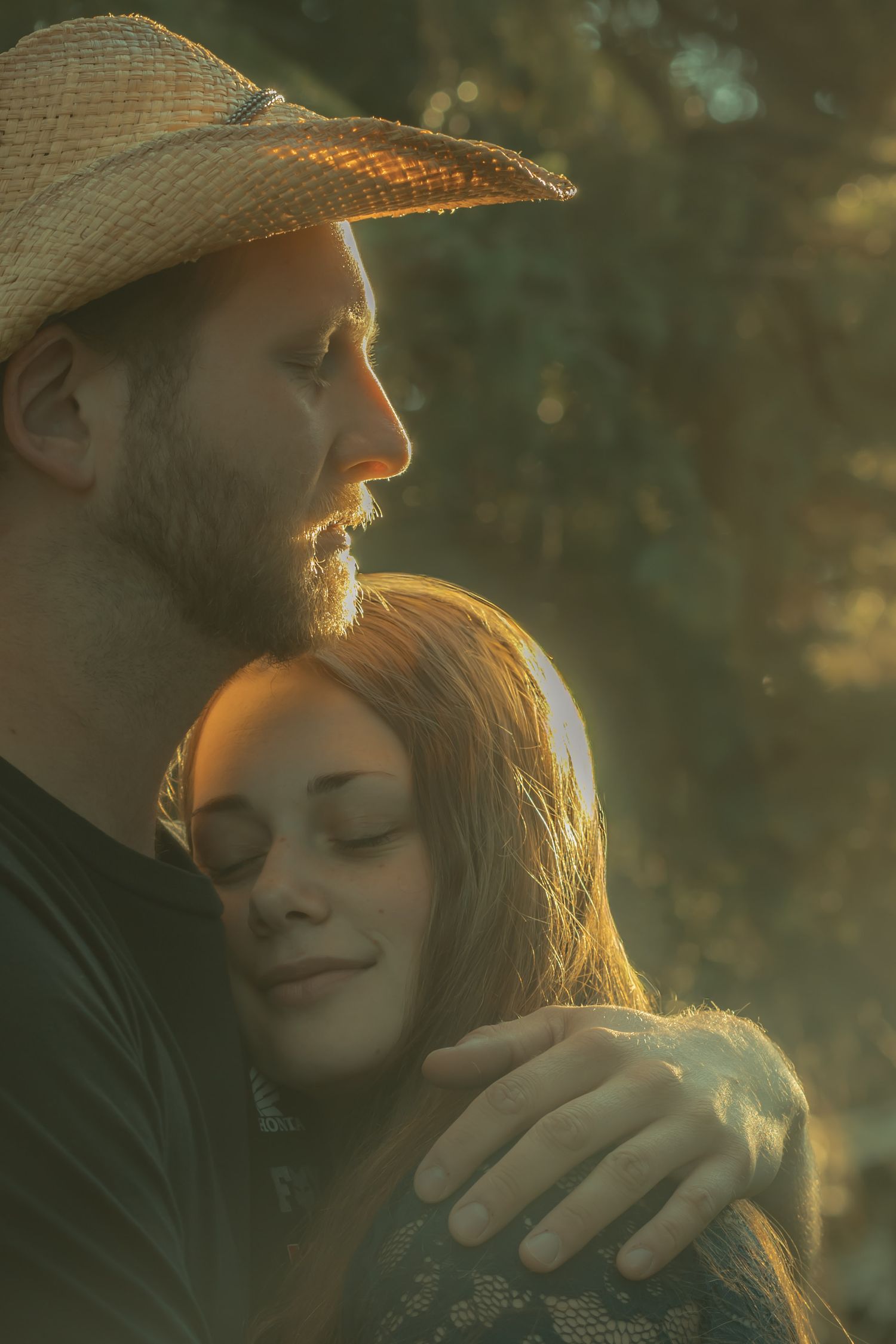 A bearded man in a straw cowboy hat tenderly embraces a red-haired woman in warm golden sunlight outdoors.