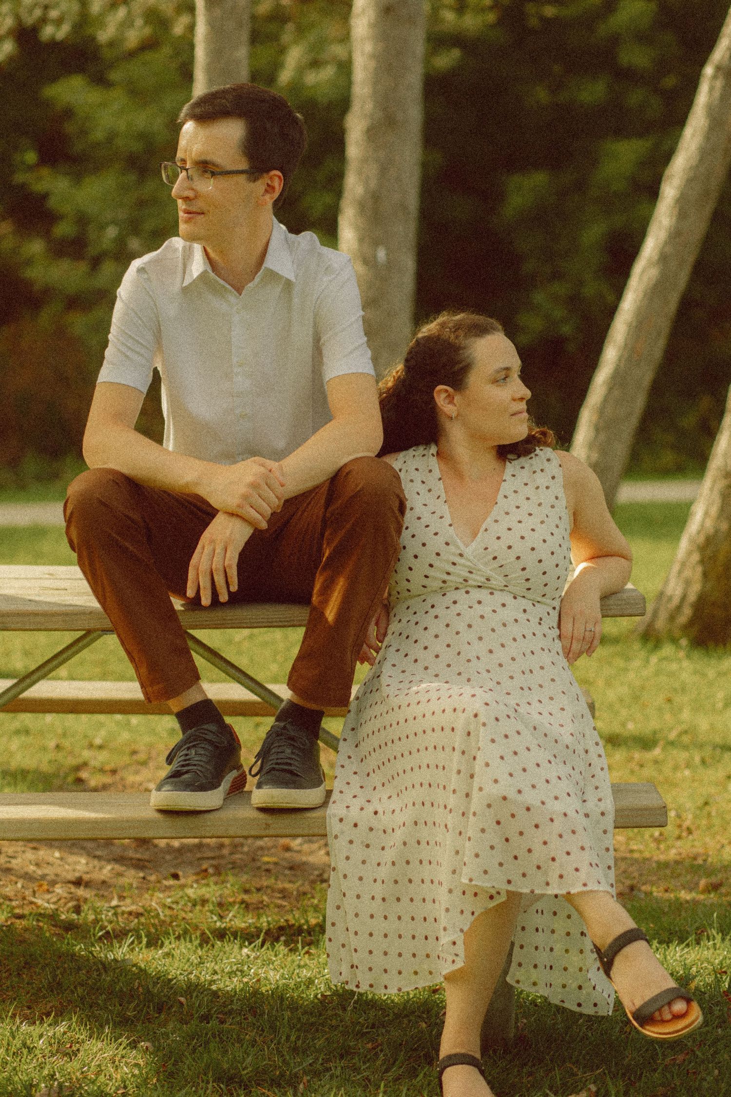 Young couple sitting on a picnic bench in a sunny park, man in brown pants and woman in polka dot dress.