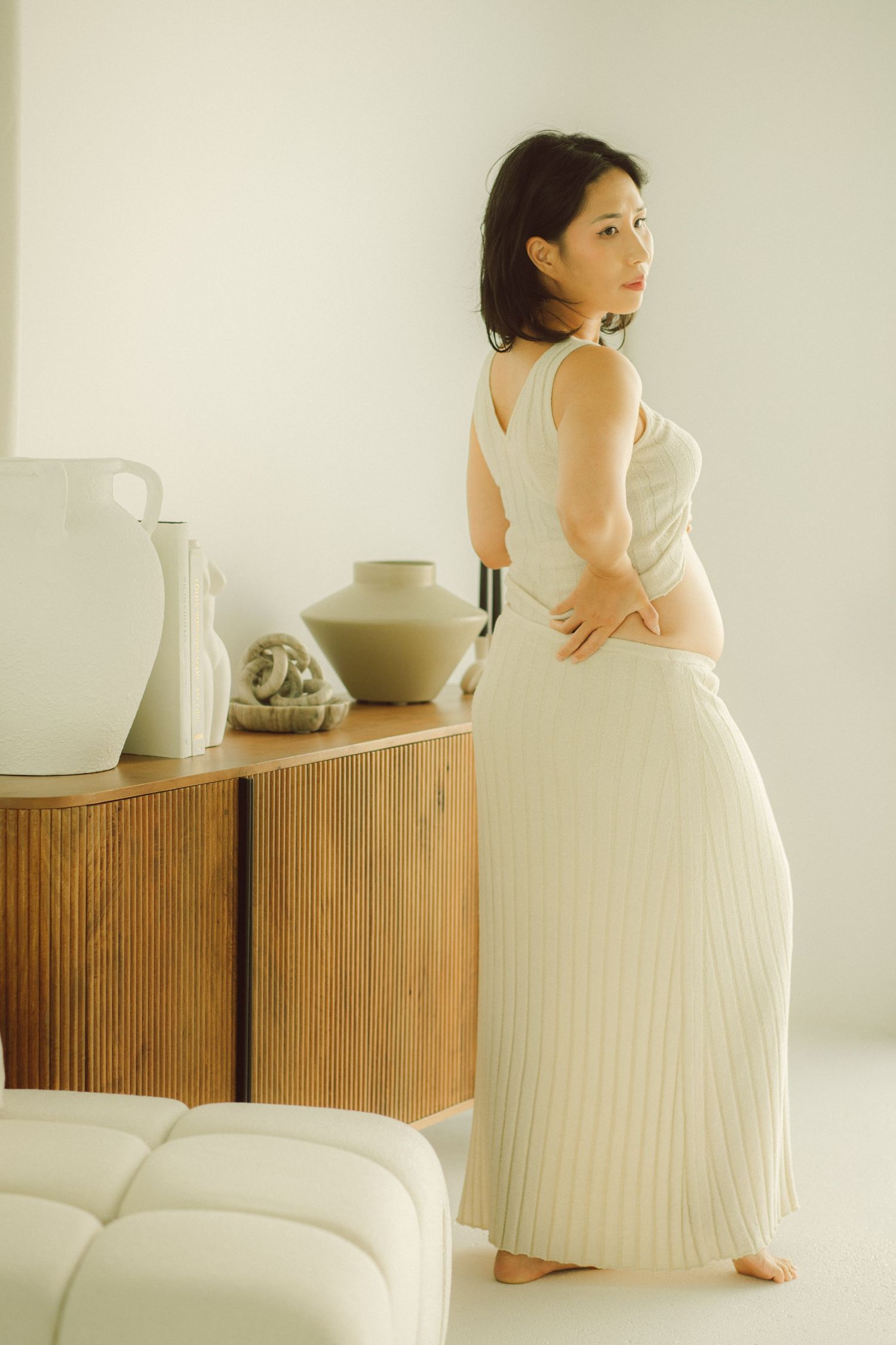 Pregnant woman in cream top and pleated skirt standing sideways near wooden dresser with ceramic vases.
