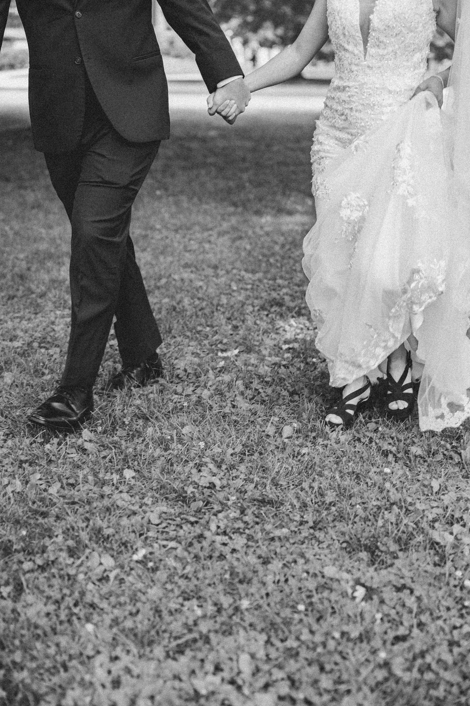 Black and white wedding photo showing close-up of bride in lace dress and groom in suit holding hands while standing on grass.