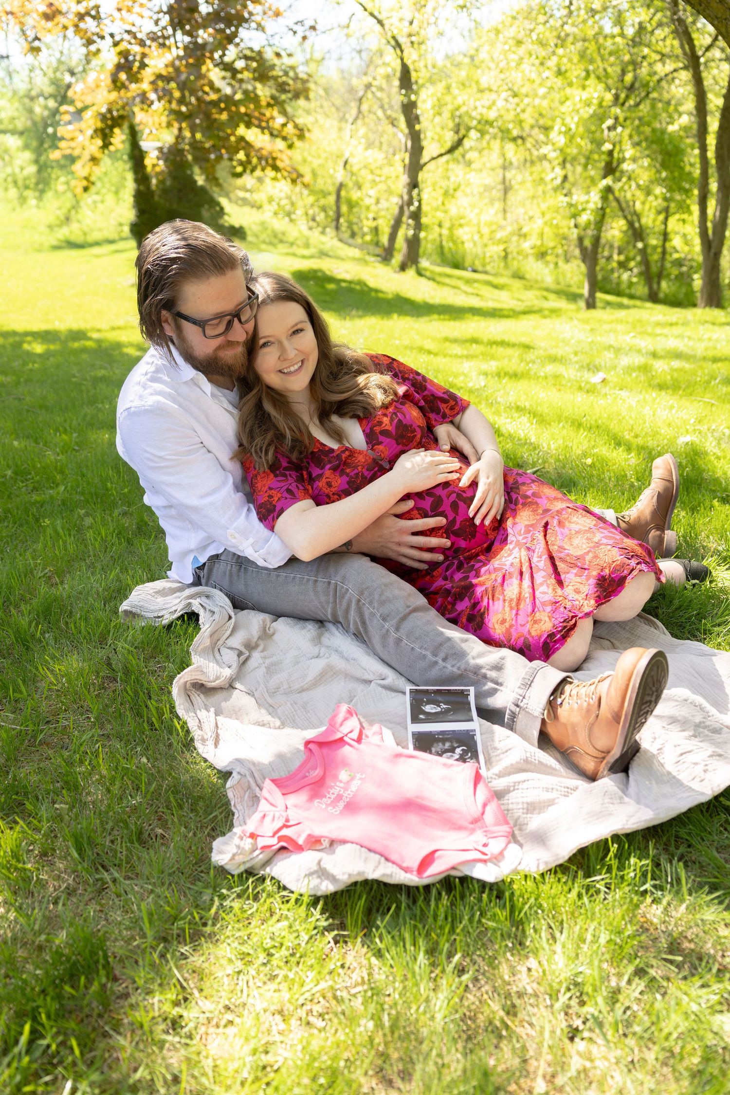 Happy pregnant couple sitting on a blanket in a sunny park with baby ultrasound photos and pink outfit.