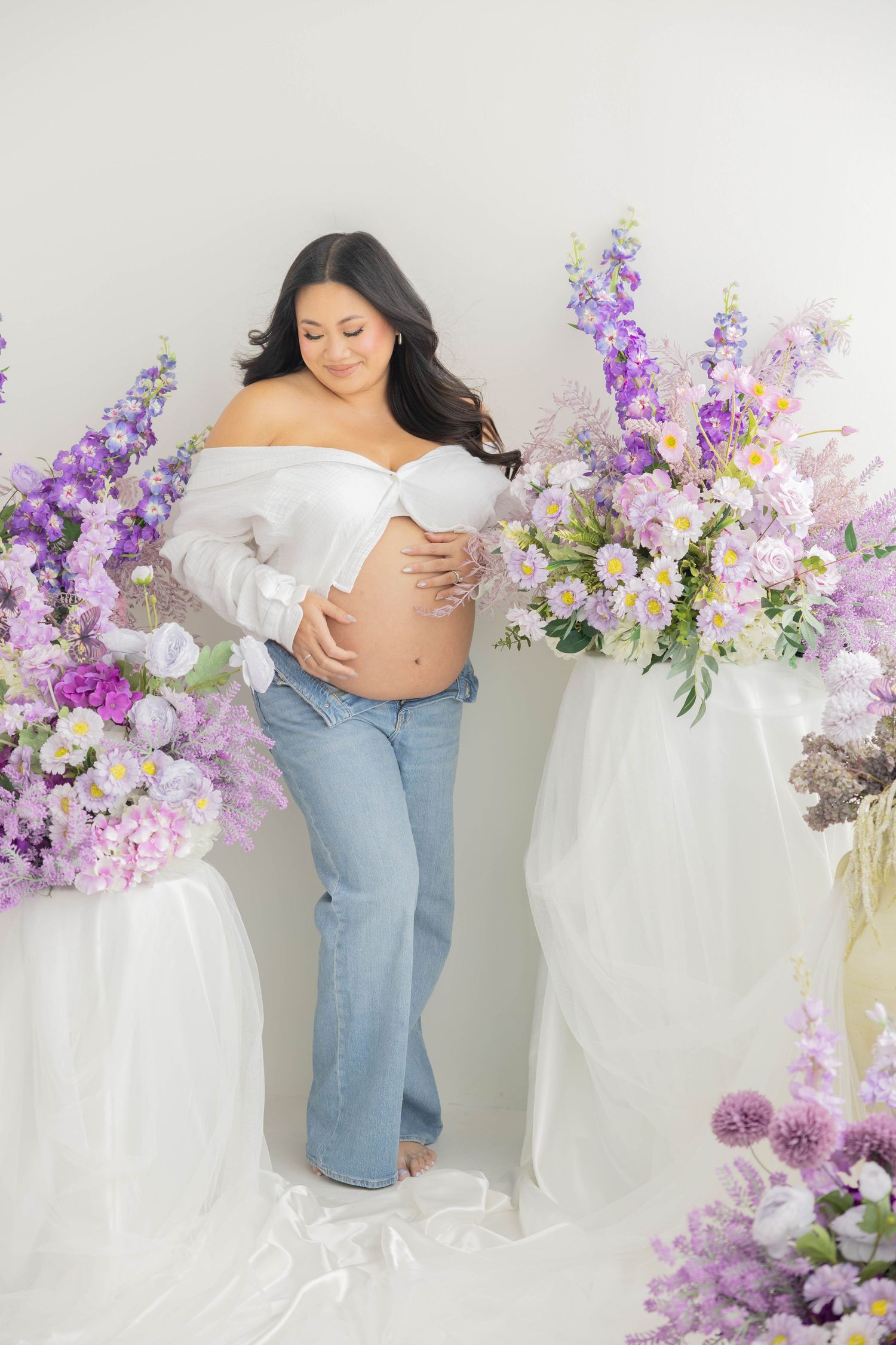 Maternity photoshoot featuring a white off-shoulder top and jeans against a backdrop with purple and pink floral arrangements.