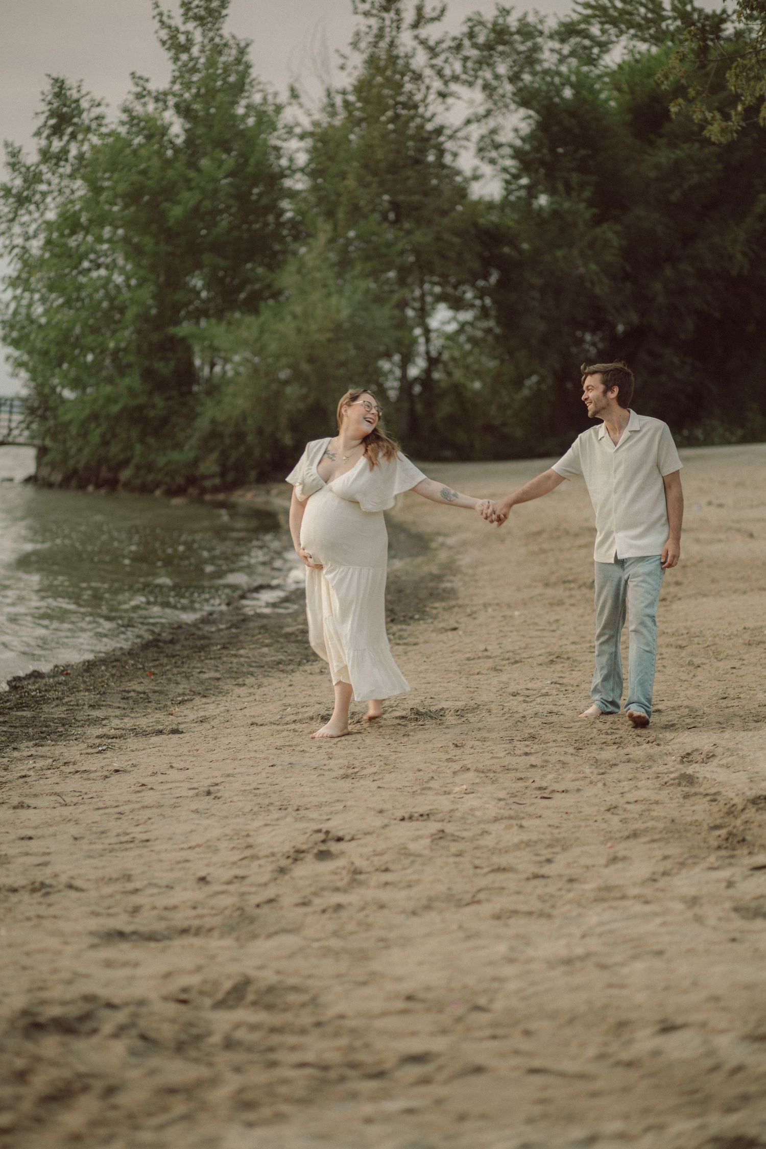 Pregnant woman in white dress holds hands with man on sandy beach, walking near green trees by the water.