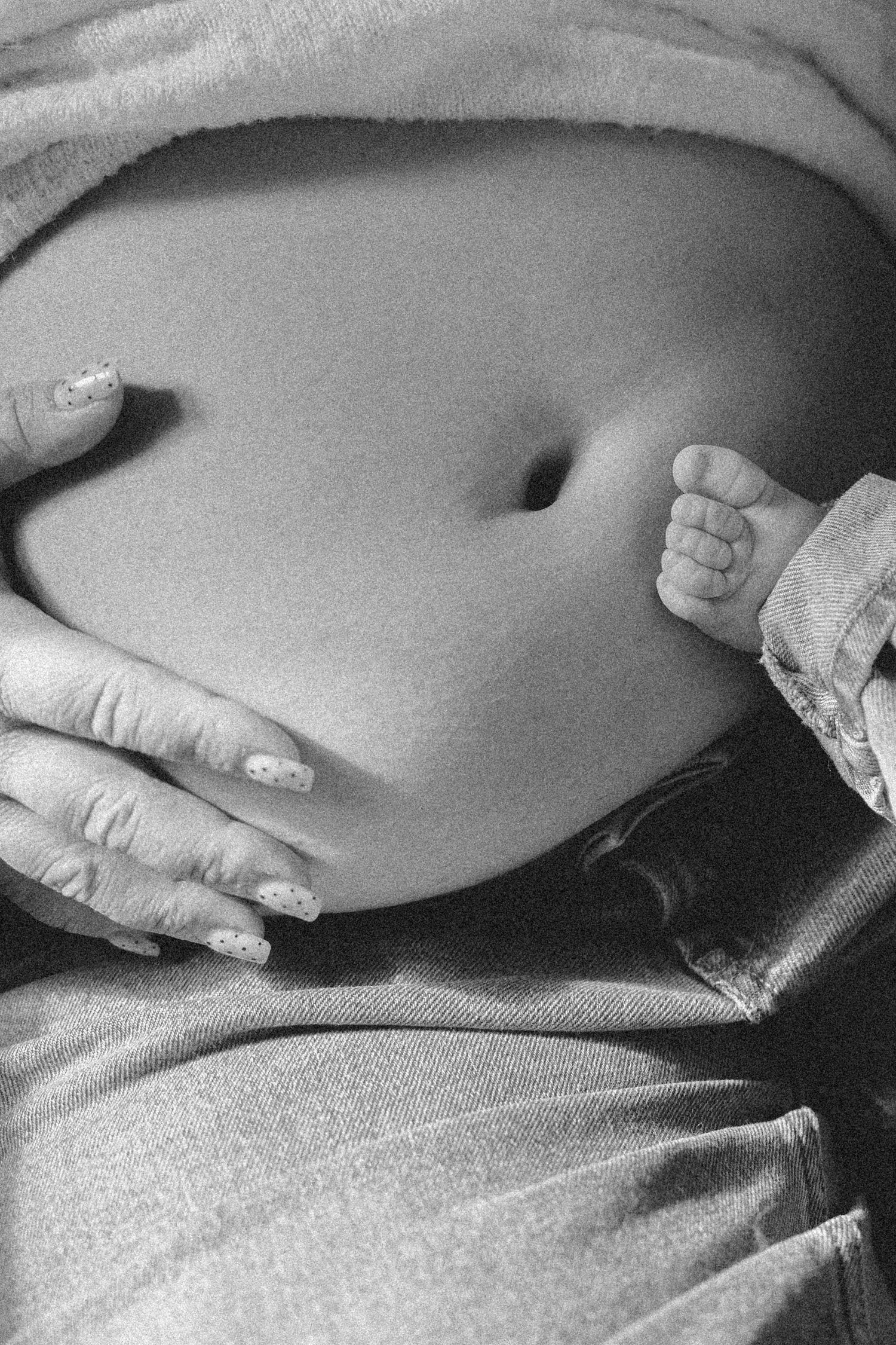 Close-up black and white image of a baby's foot and hands resting on a bare pregnant belly.