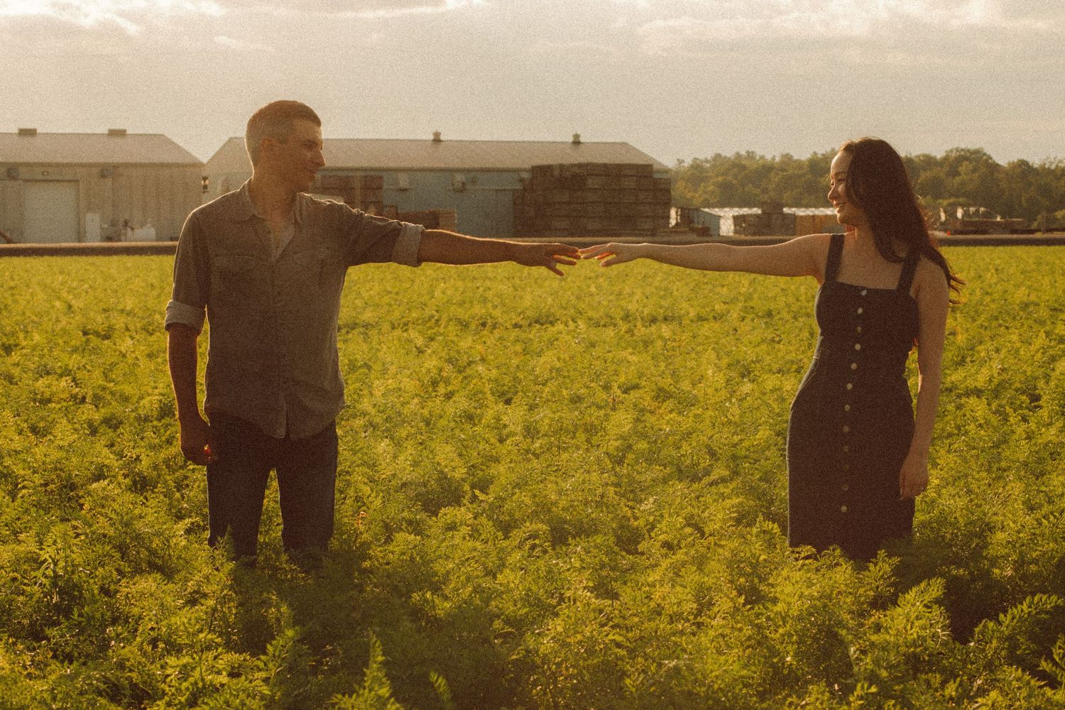 A man and woman reach toward each other across a lush green field at golden hour near farm buildings.